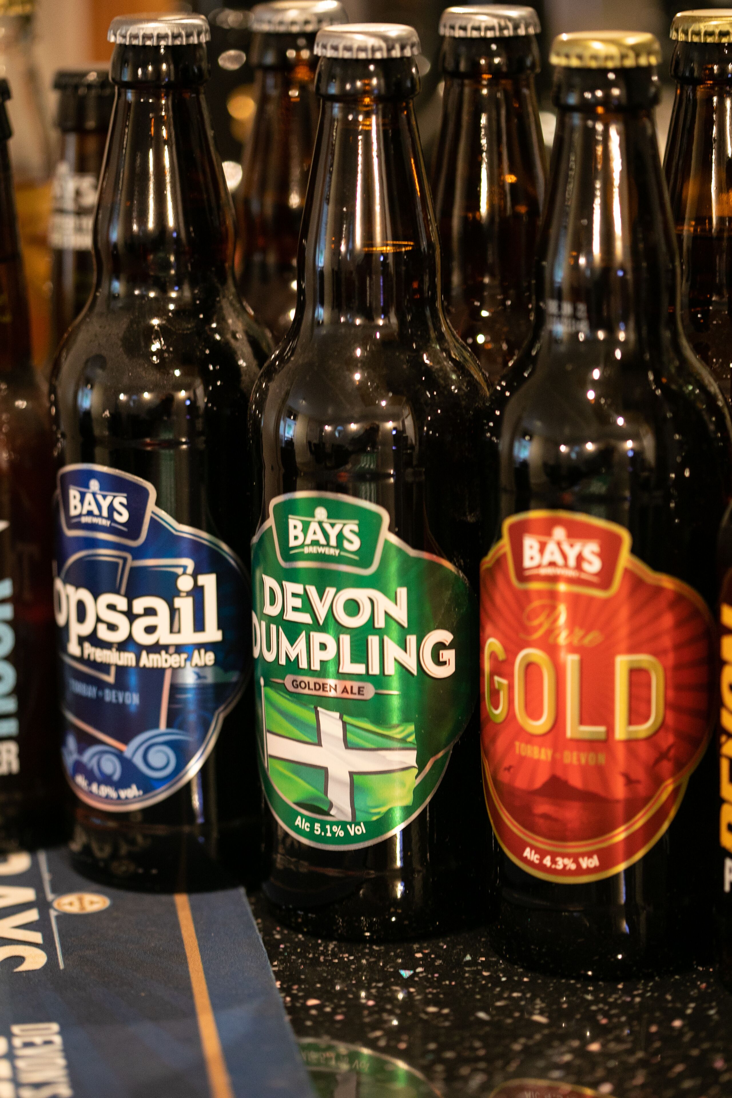 Eine Reihe Bierflaschen mit Etiketten, darunter 'Bays', 'Devon Dumpling' und 'Gold'.