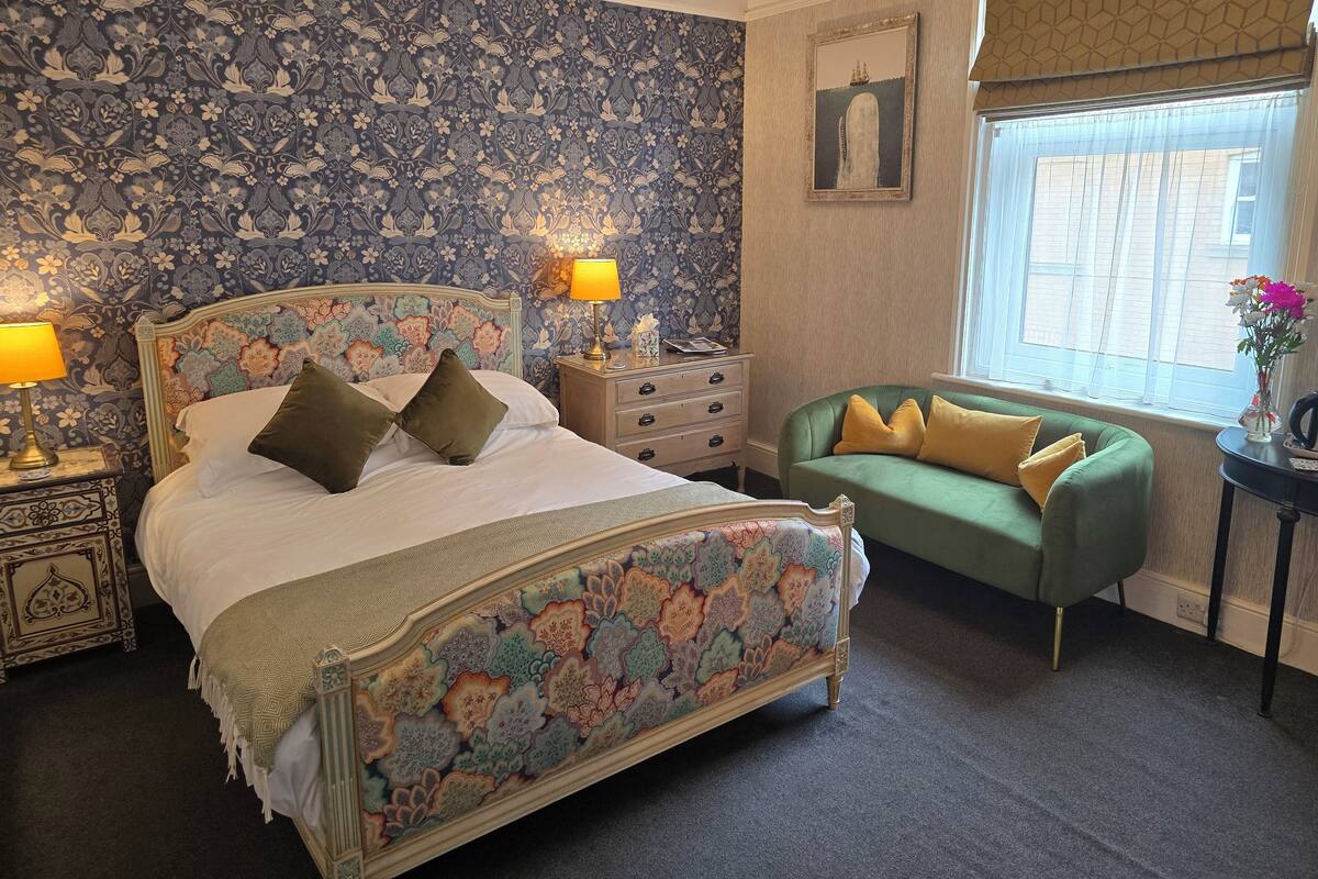 A bedroom with a floral-patterned bed, a green sofa, a nightstand with a lamp, and a window with a view of buildings.