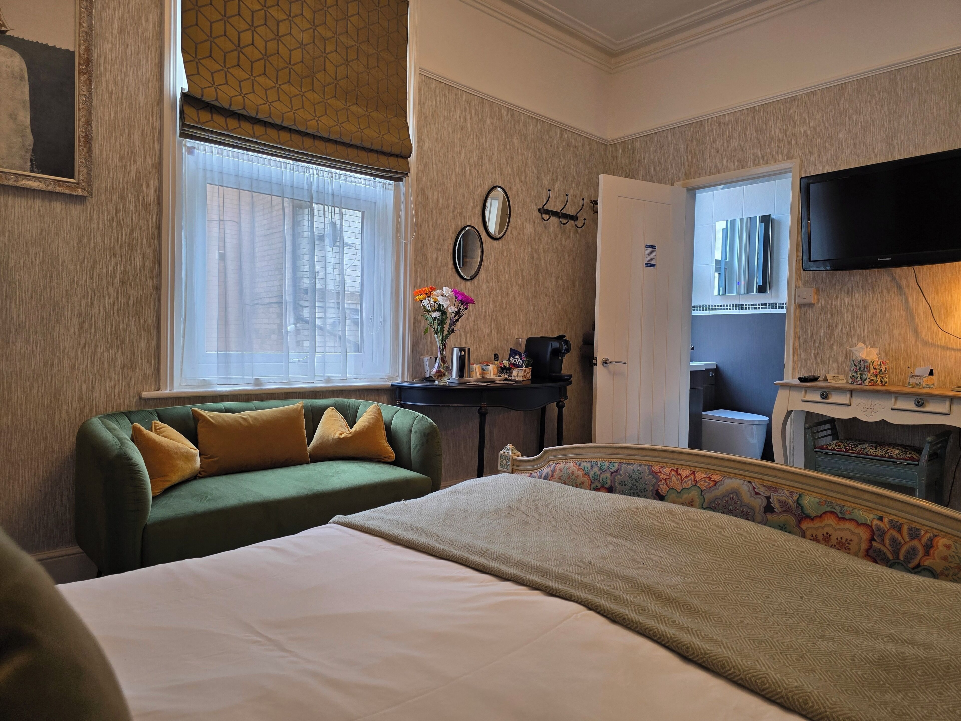 A hotel room with a green sofa, a bed with a patterned headboard, a desk with a coffee maker, a mirror, and a television.