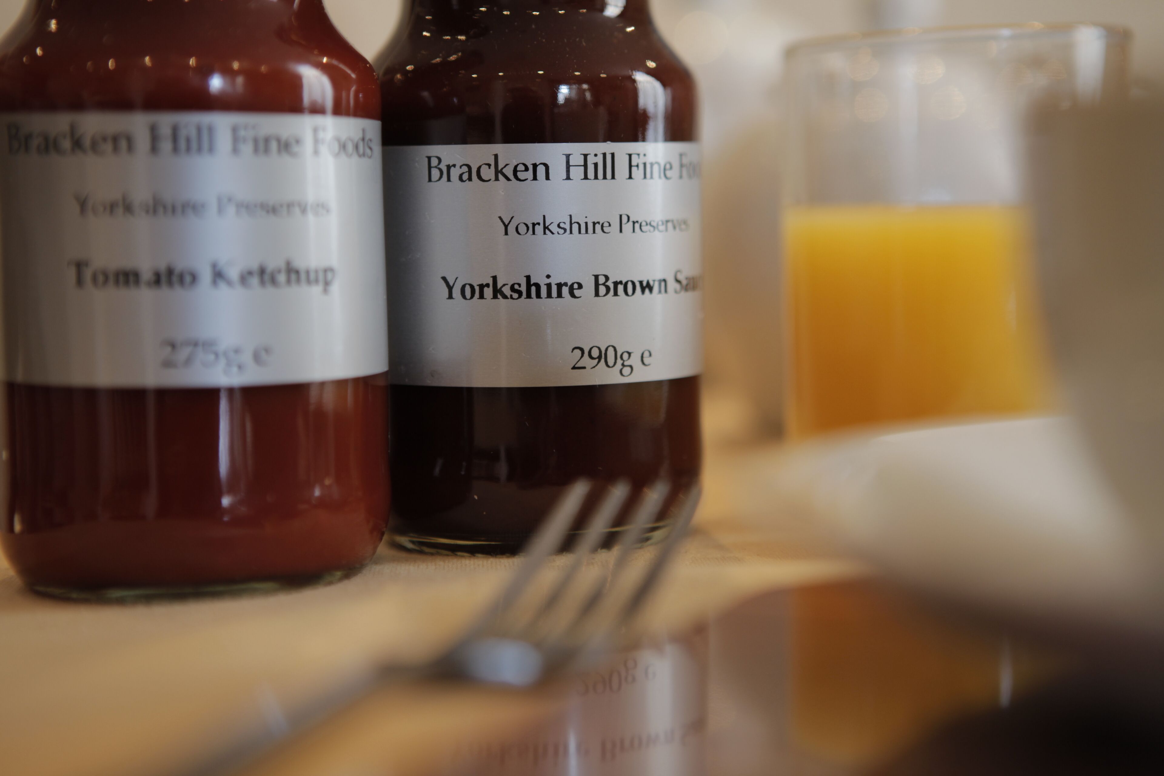 Two bottles of Bracken Hill Fine Foods, one labeled Tomato Ketchup and the other Yorkshire Brown Sauce, with a glass of orange juice in the background.