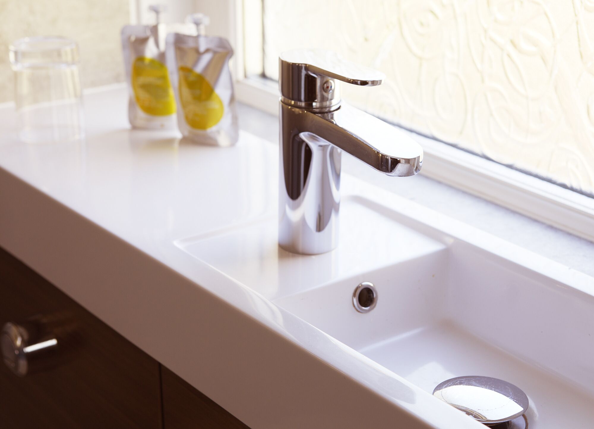 Un lavabo moderne pour la salle de bain, doté d’un robinet chromé, une fenêtre ornée d’un papier peint décoratif et deux bouteilles en verre contenant un liquide jaune.