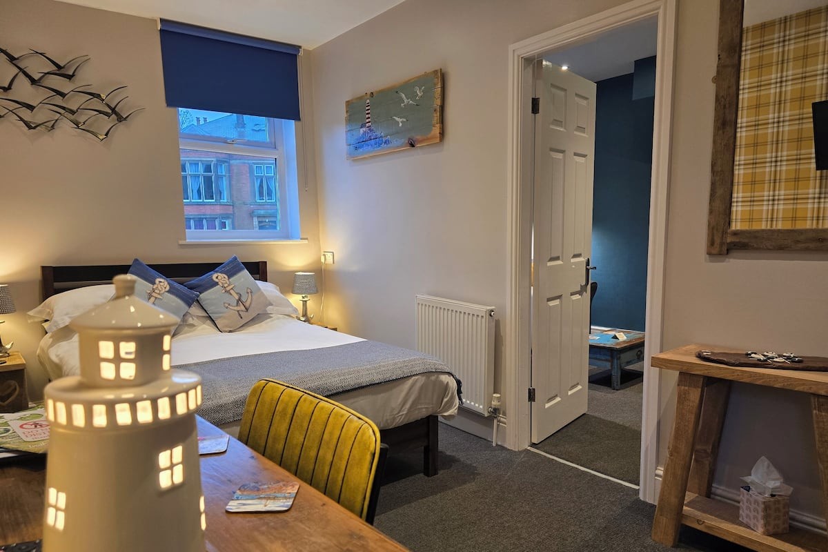 A bedroom with a bed, bedside lamp, and a nightstand. There is a window with a view of buildings outside.