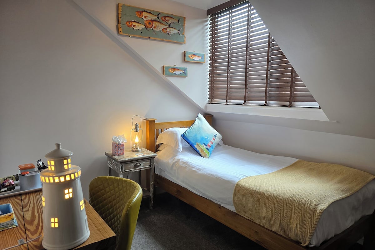 A cozy bedroom with a bed, a nightstand, a lamp, a chair, and a window with blinds.