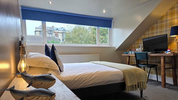 A bedroom with a bed, a desk with a computer, a lamp, and a view of buildings outside. - Ashburton House (Scarborough)