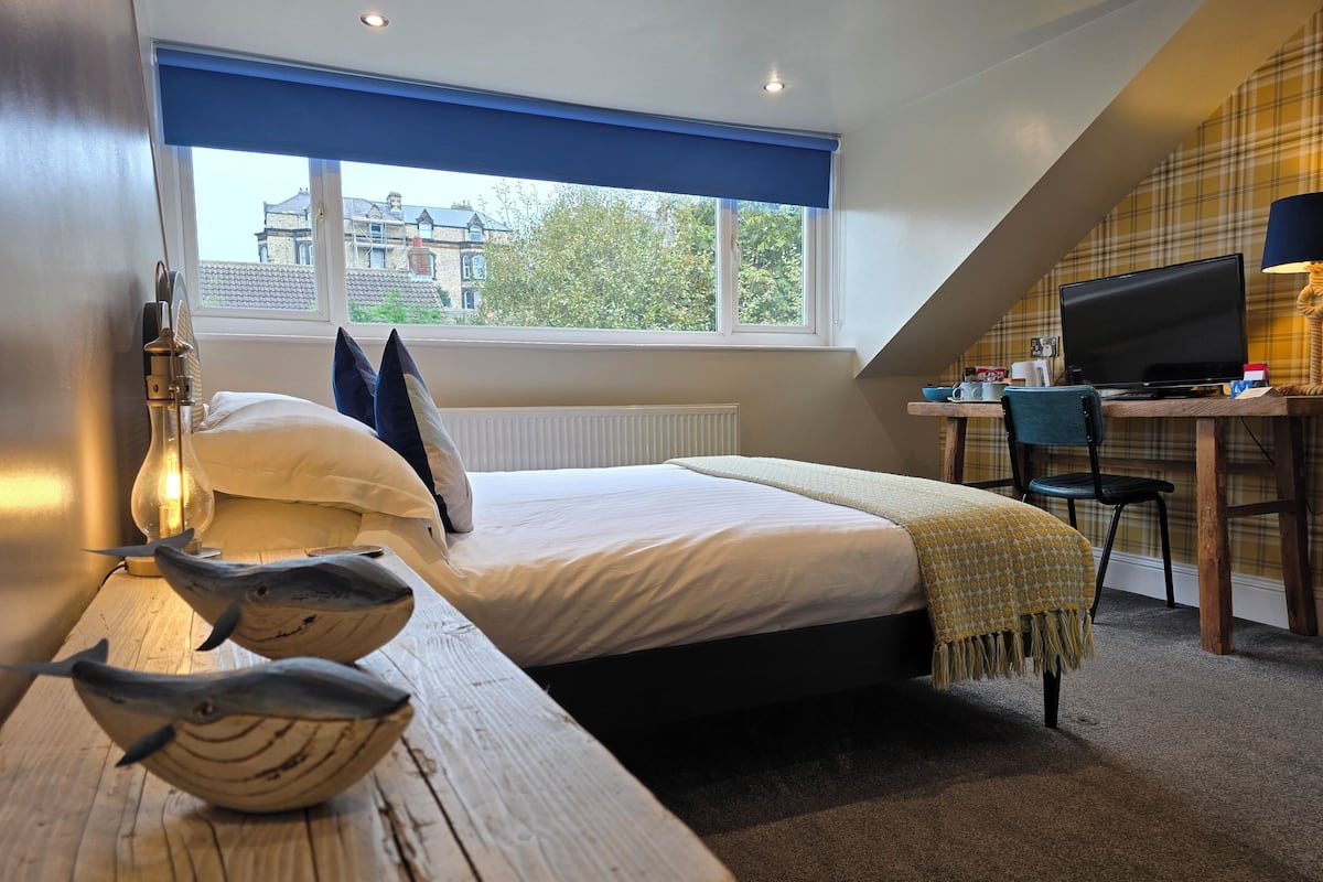 A bedroom with a bed, a desk with a computer, a lamp, and a view of buildings outside.