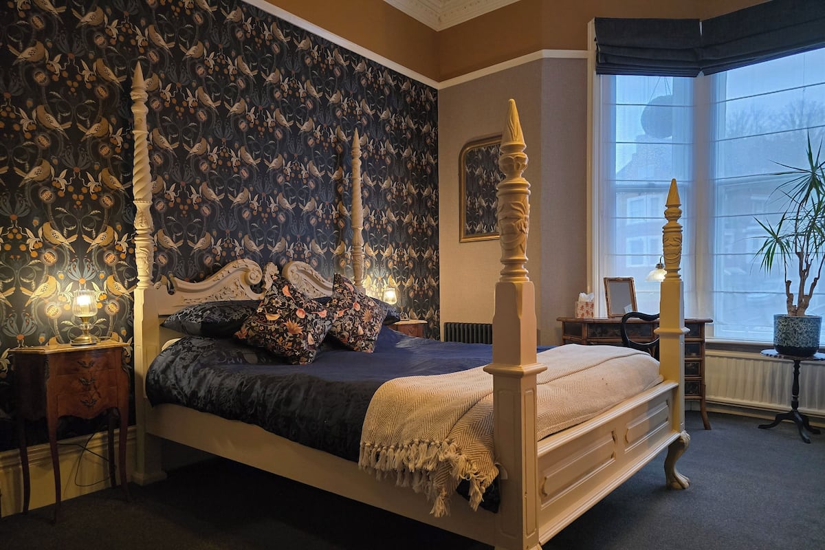 A four-poster bed with ornate carvings, a dark patterned wallpaper, and a large window with a view of the outside.