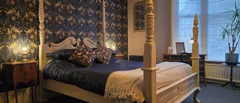 Una cama con dosel y elaboradas tallas, un papel tapiz oscuro con estampado y un ventanal con vistas al exterior.