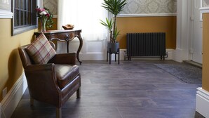 A room with a brown leather sofa, a plaid cushion, a small table with a vase of flowers, a potted plant, and a radiator. - Ashburton House (Scarborough)