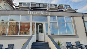 Un edificio con el nombre 'Brooklyn' en la fachada, que cuenta con una escalera que conduce a la entrada, asientos exteriores con sillas y plantas en macetas.