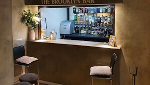 Zona de bar con una barra que exhibe varias botellas y un televisor montado en la parte superior. Hay dos taburetes y una silla.
