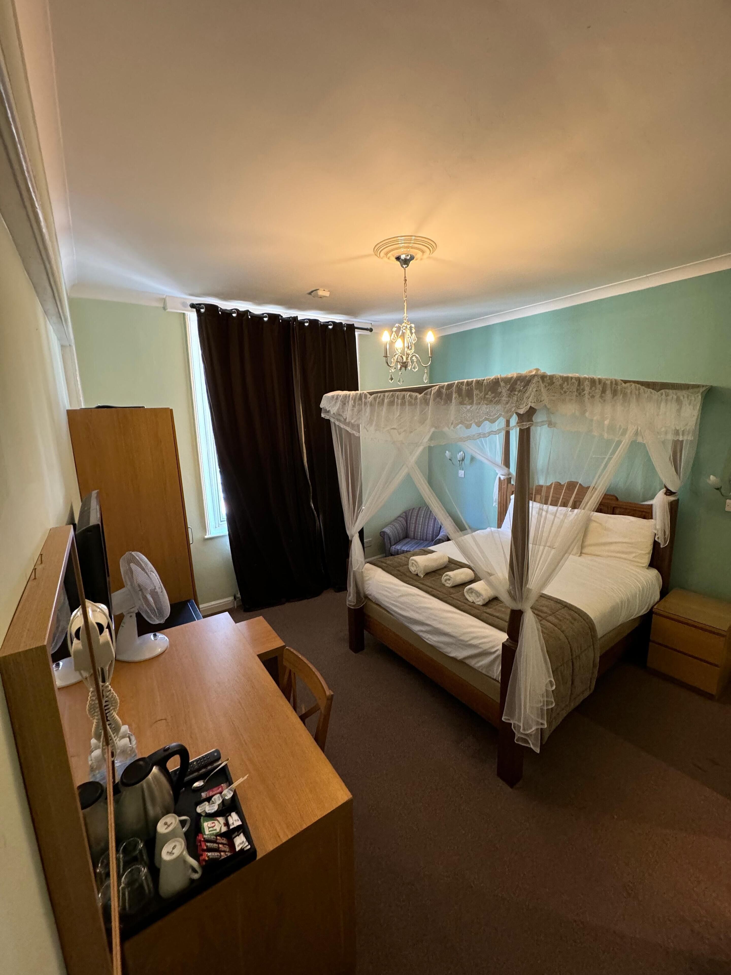 A hotel room with a four-poster bed, a desk with a fan, a chair, a bedside table, and a nightstand.
