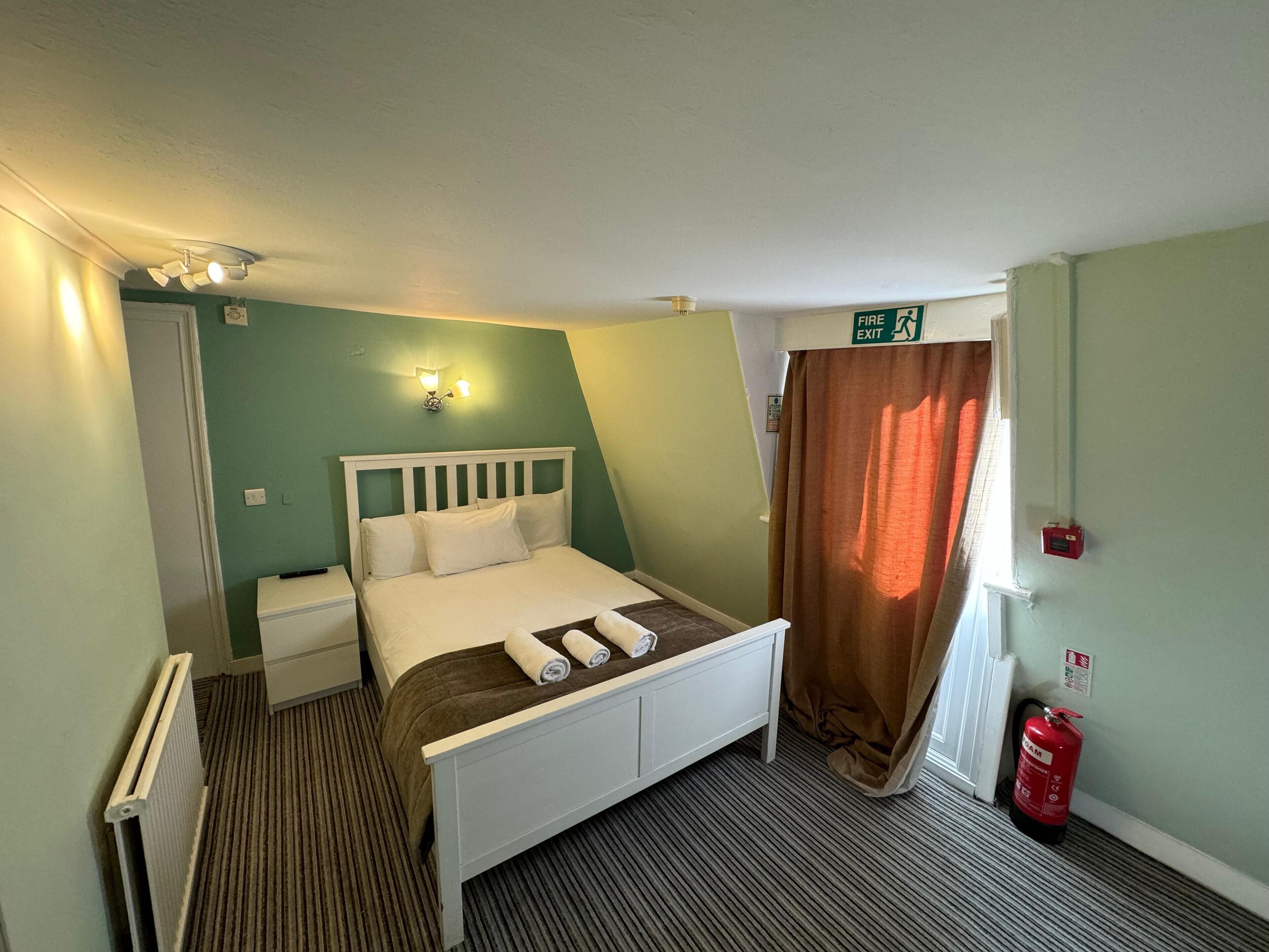 A single-bed room with a white headboard, a bedside table, a radiator, a brown curtain, a fire exit sign, and a fire extinguisher.