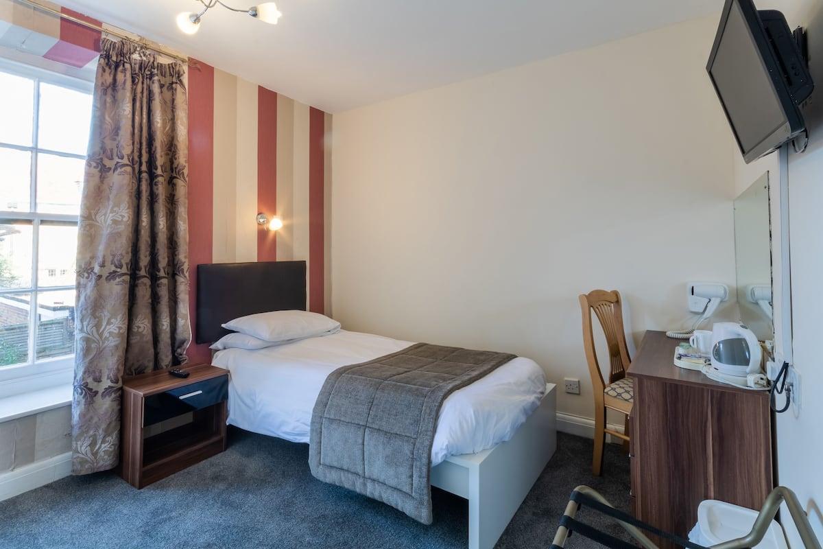 A hotel room with a bed, a bedside table, a chair, a desk with a mirror, and a TV.