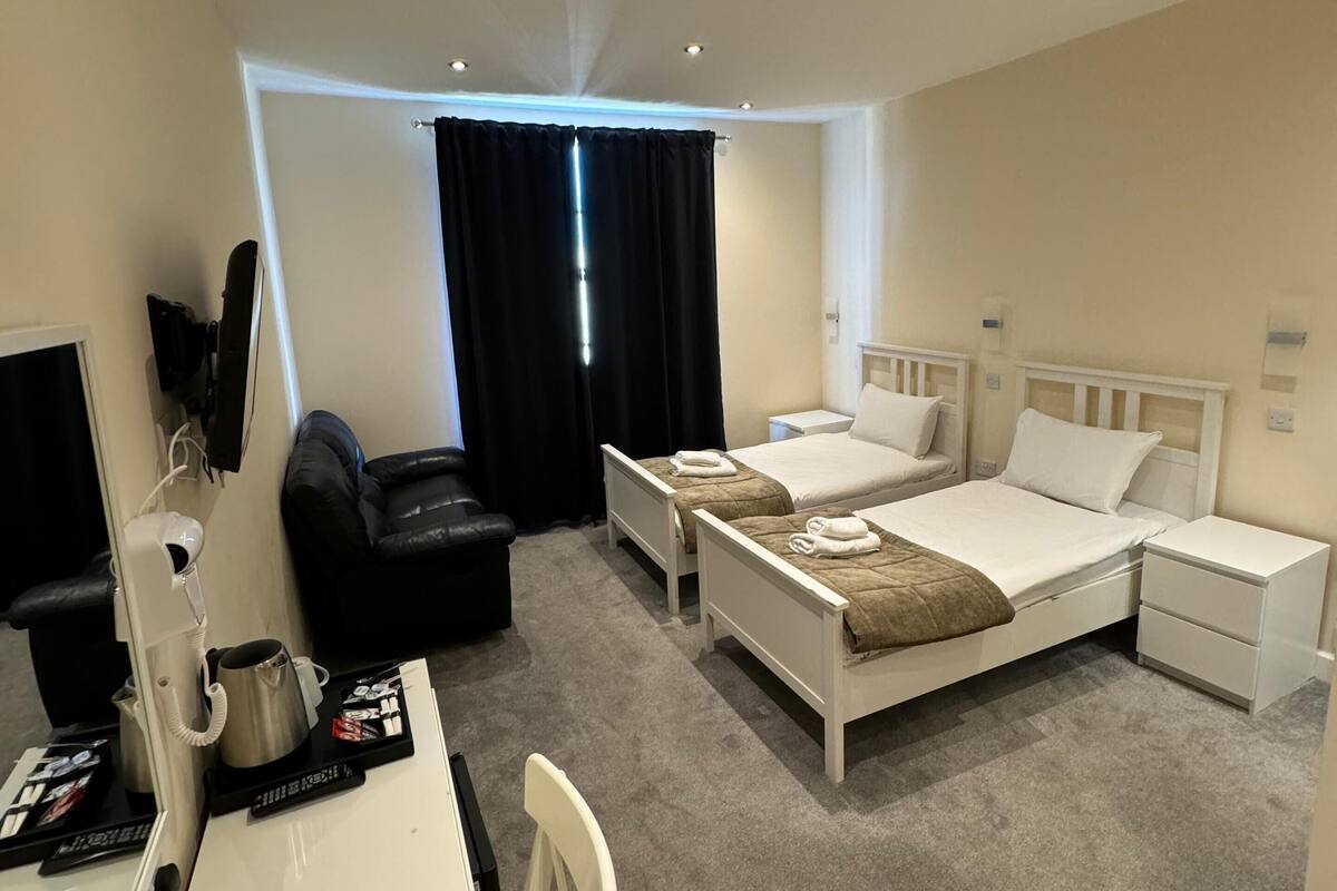A hotel room with two single beds, a desk with a chair, a black leather sofa, a small table with a kettle and a cup, and a TV.