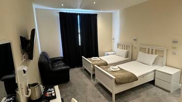 A hotel room with two single beds, a desk with a chair, a black leather sofa, a small table with a kettle and a cup, and a TV.