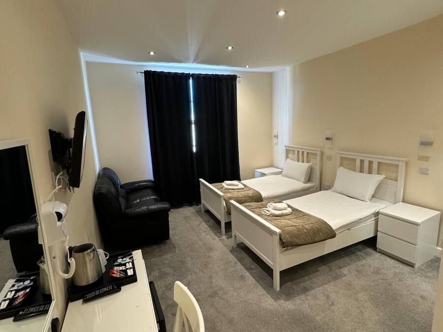 A hotel room with two single beds, a desk with a chair, a black leather sofa, a small table with a kettle and a cup, and a TV.