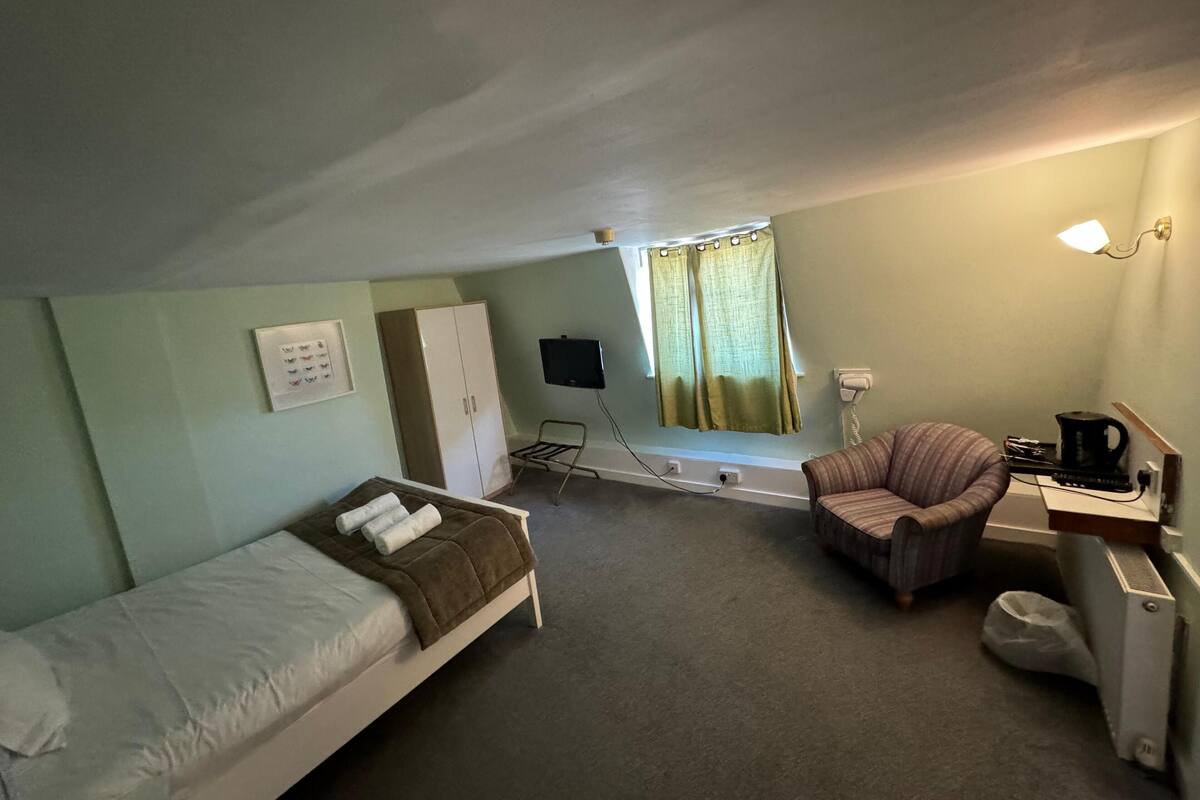 A small, single-room hotel room with a bed, a chair, a desk with a computer, a television, and a window with curtains.