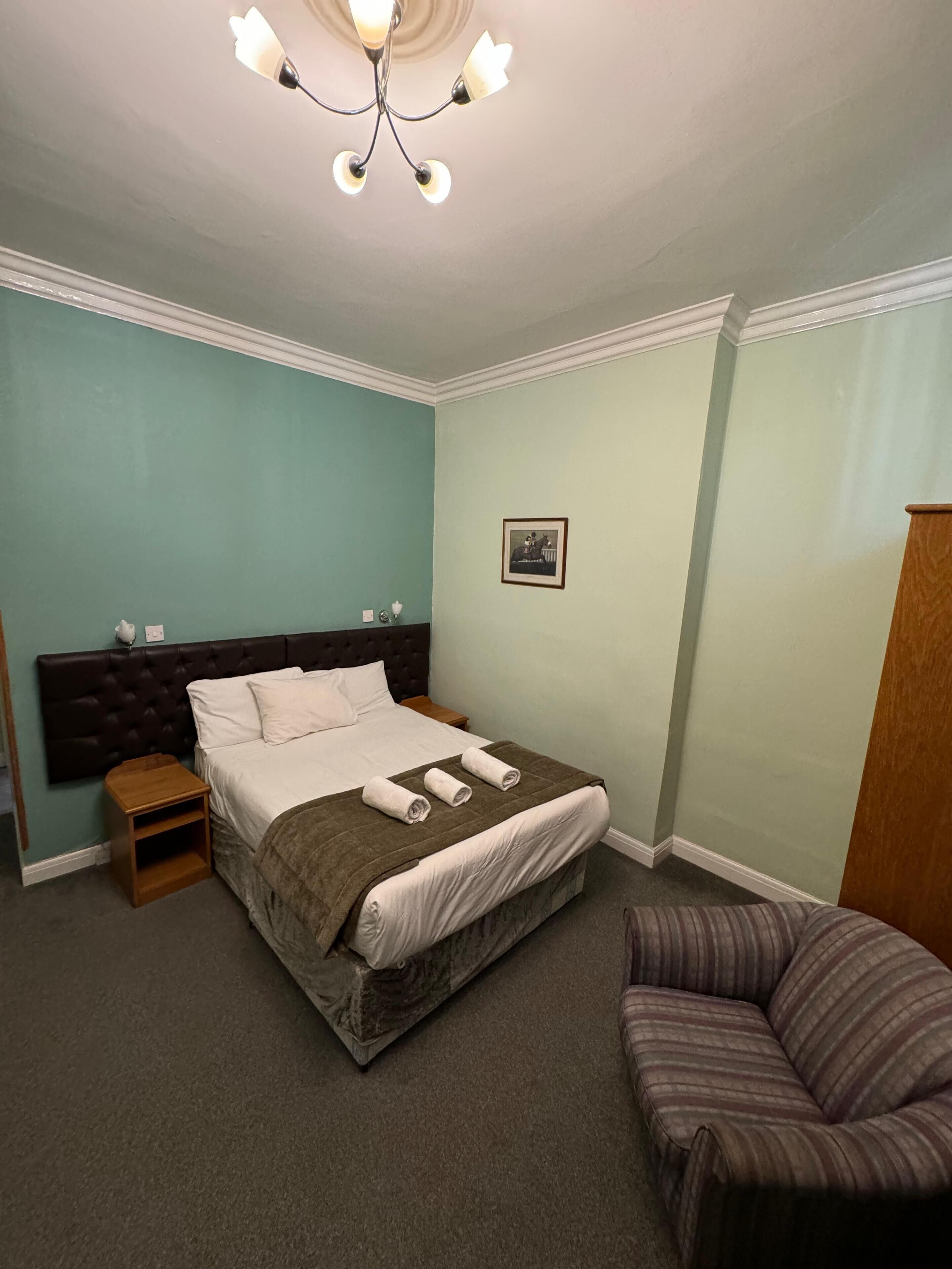 A neatly made bed with a dark headboard, a small bedside table, a chair, and a picture on the wall.