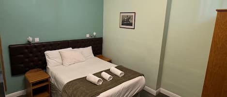 A neatly made bed with a dark headboard, a small bedside table, a chair, and a picture on the wall.