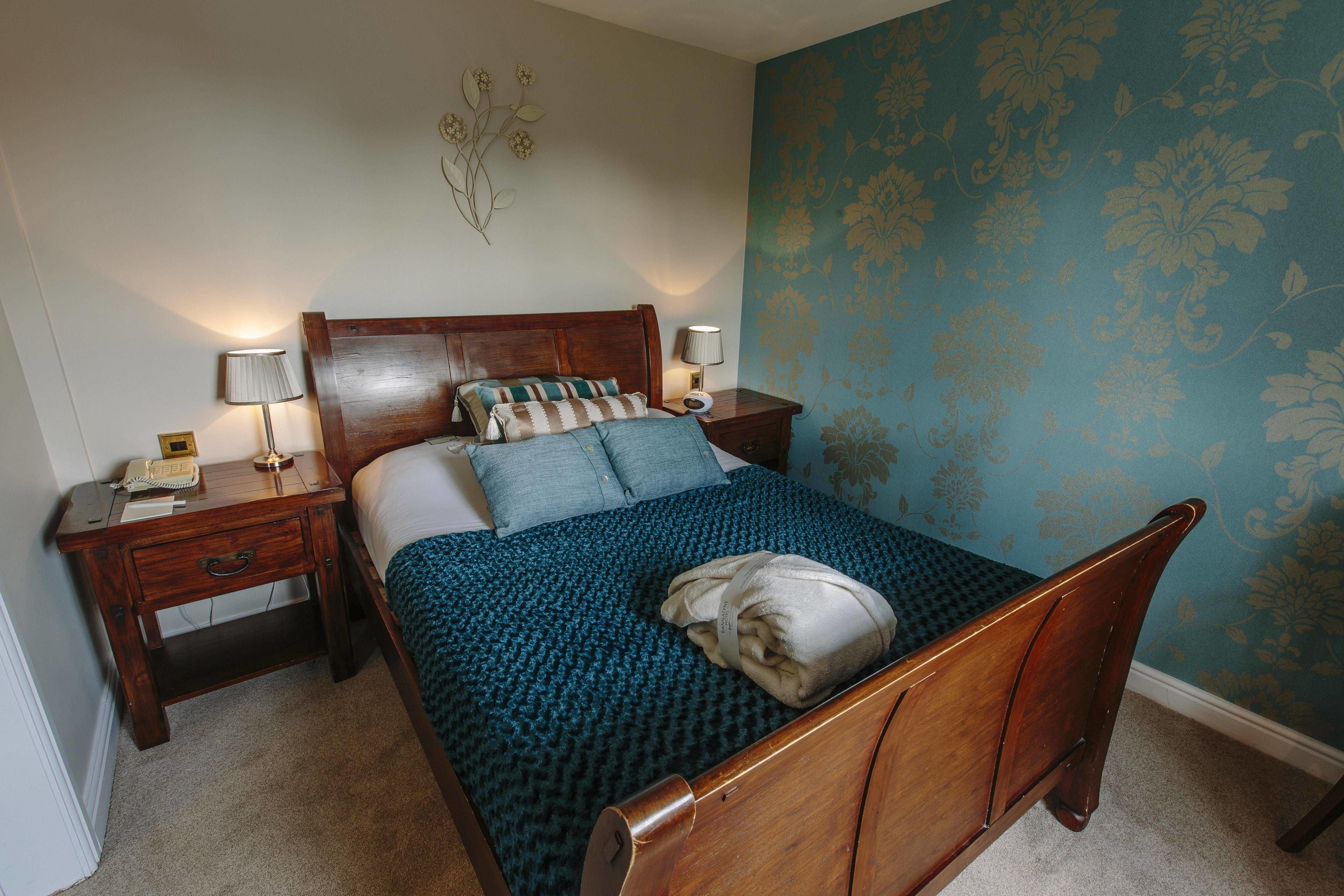 A neatly made bed with a blue patterned bedspread and pillows, a wooden headboard, and a nightstand with a lamp and a telephone.