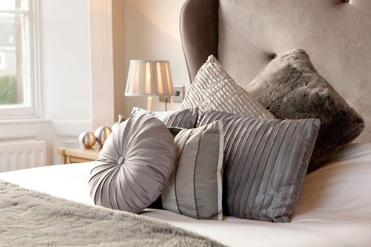 Una cama bien hecha con un cabecero capitonado, varios almohadones y una manta mullida.