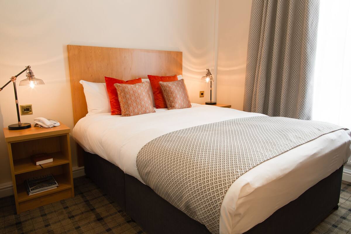 Una cama bien hecha con una colcha estampada, dos lámparas de noche, una mesita de noche con un teléfono y libros, y una ventana con cortinas.