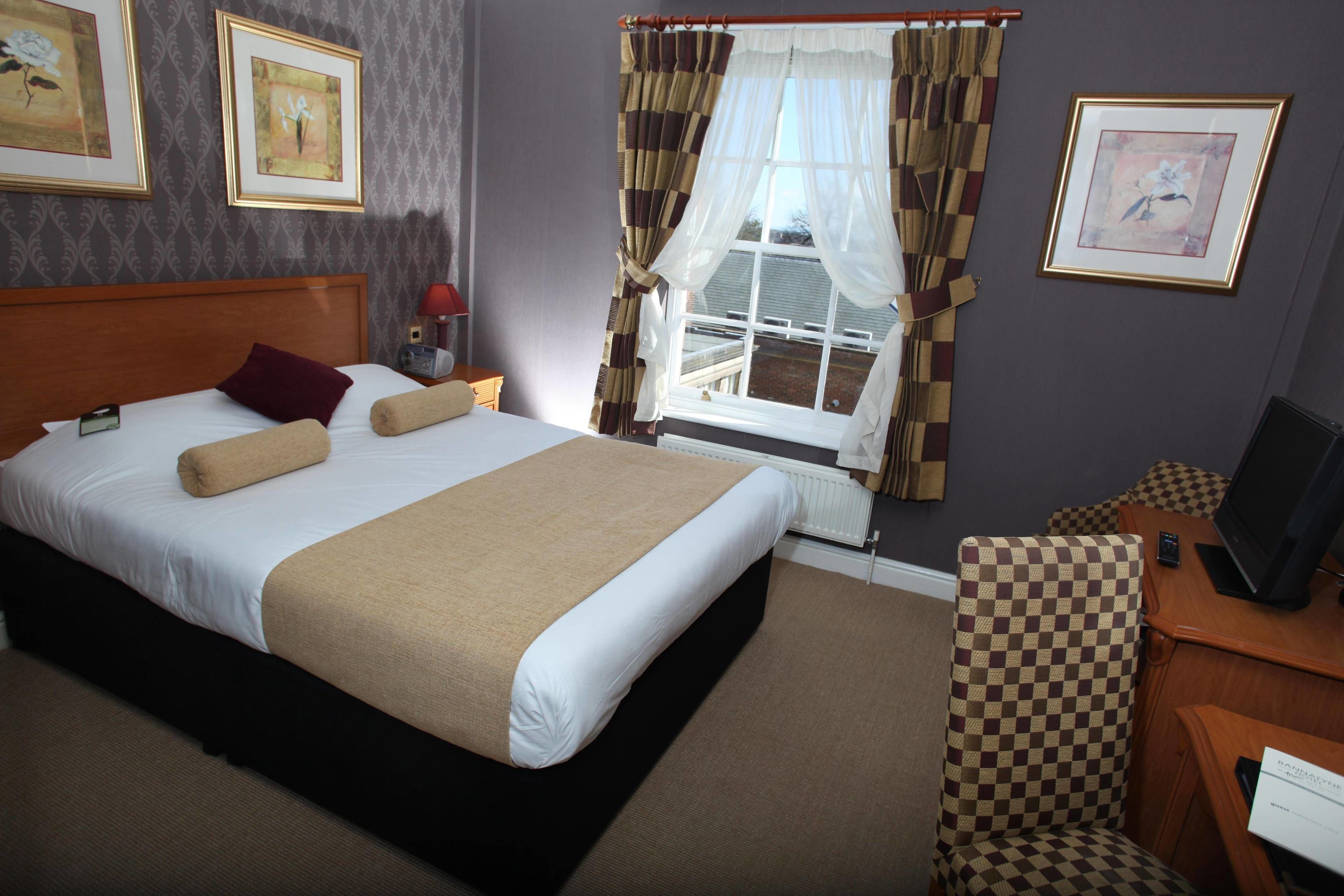 A hotel room with a large bed, a chair, a desk with a computer, and a window with curtains.