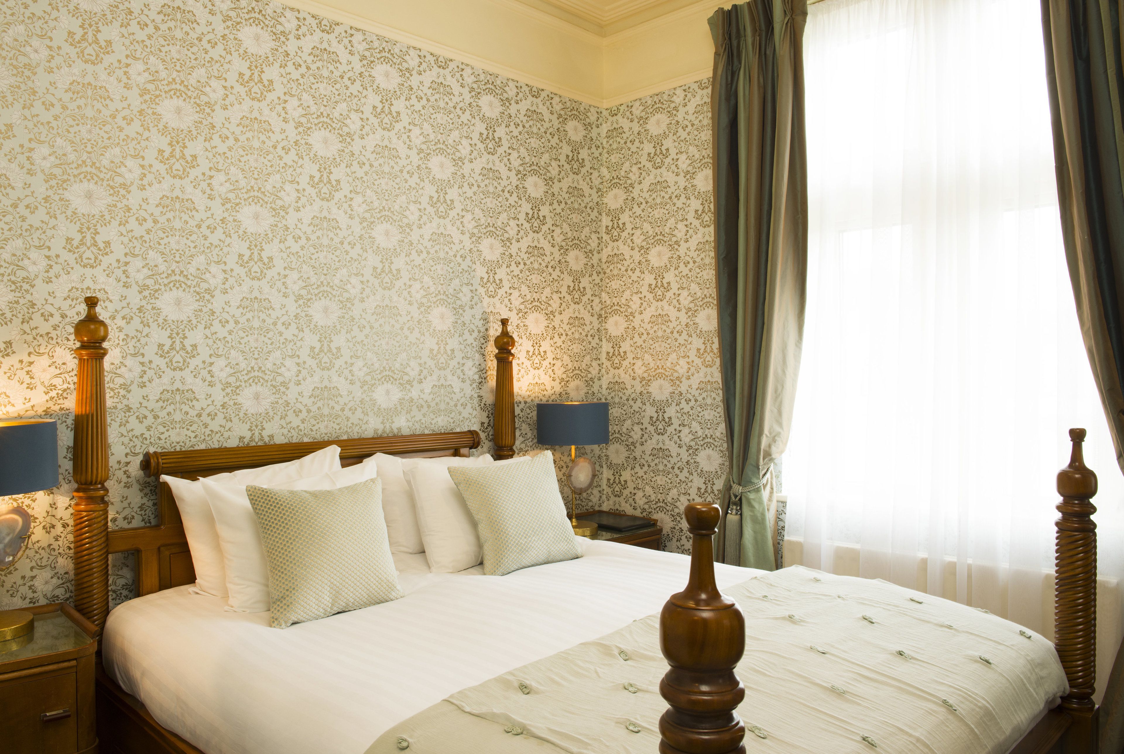 A classic four-poster bed with a wooden headboard, flanked by two bedside tables with lamps, in a room with floral wallpaper and a large window with curtains.