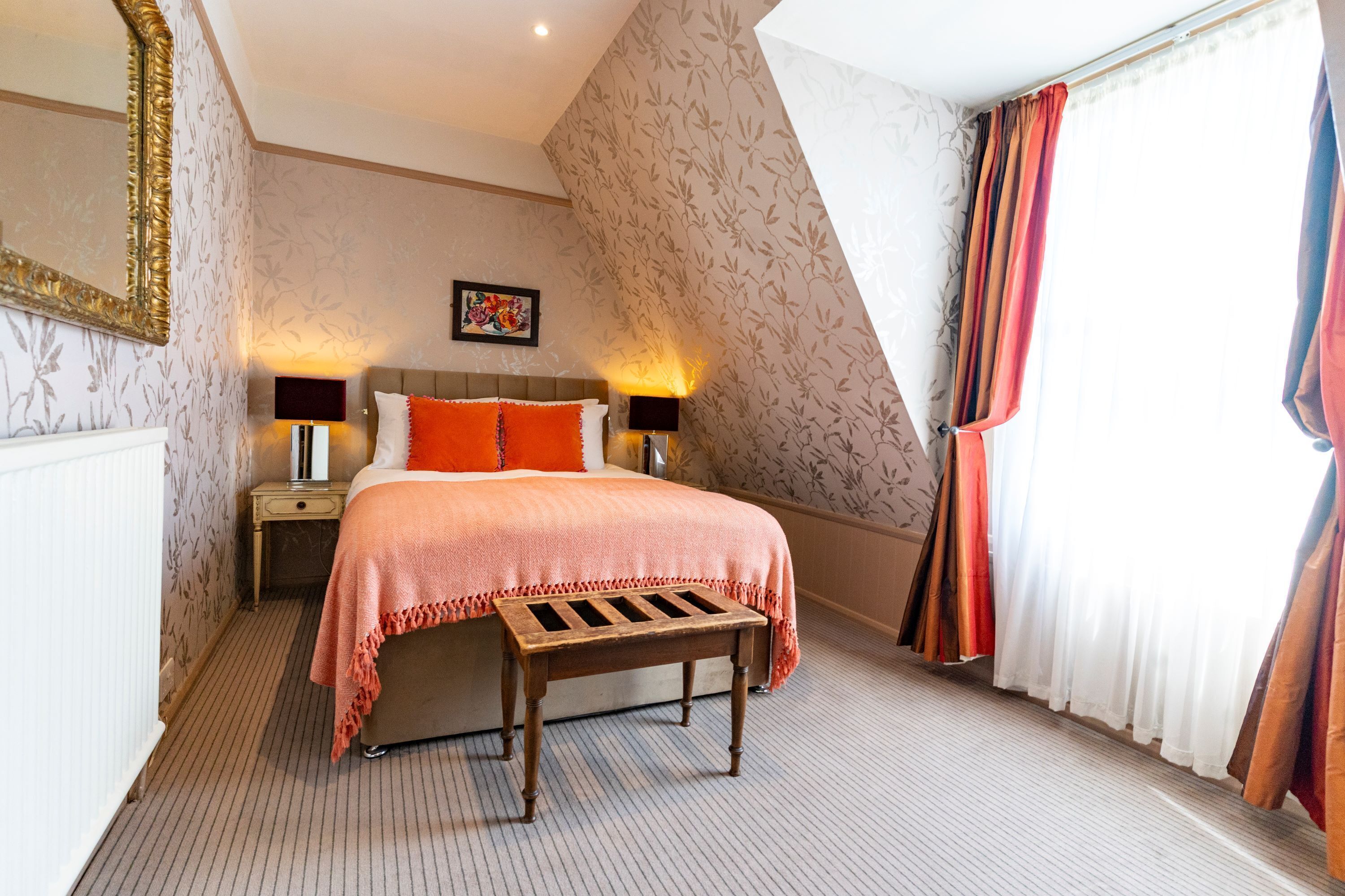A bedroom with a bed, nightstands, a bench, a mirror, and a window with curtains.