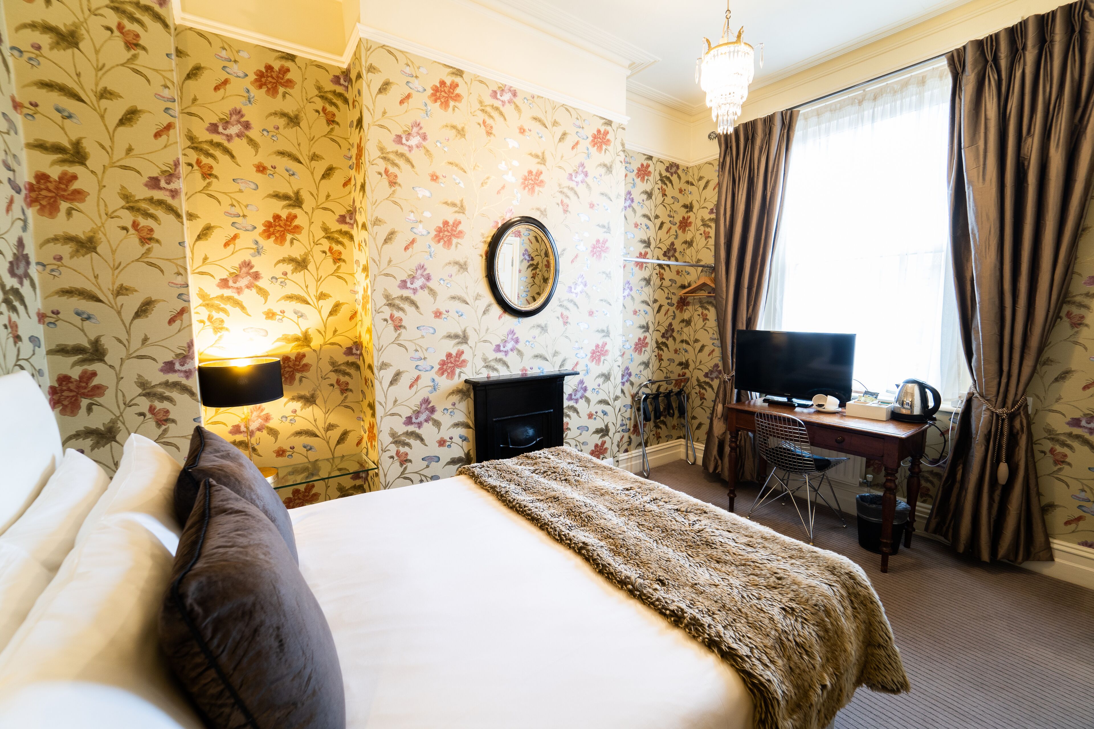 Una habitación de hotel con papel tapiz floral, una cama con cubrecama blanco y manta marrón, un escritorio con computadora y una silla, y una ventana con cortinas.