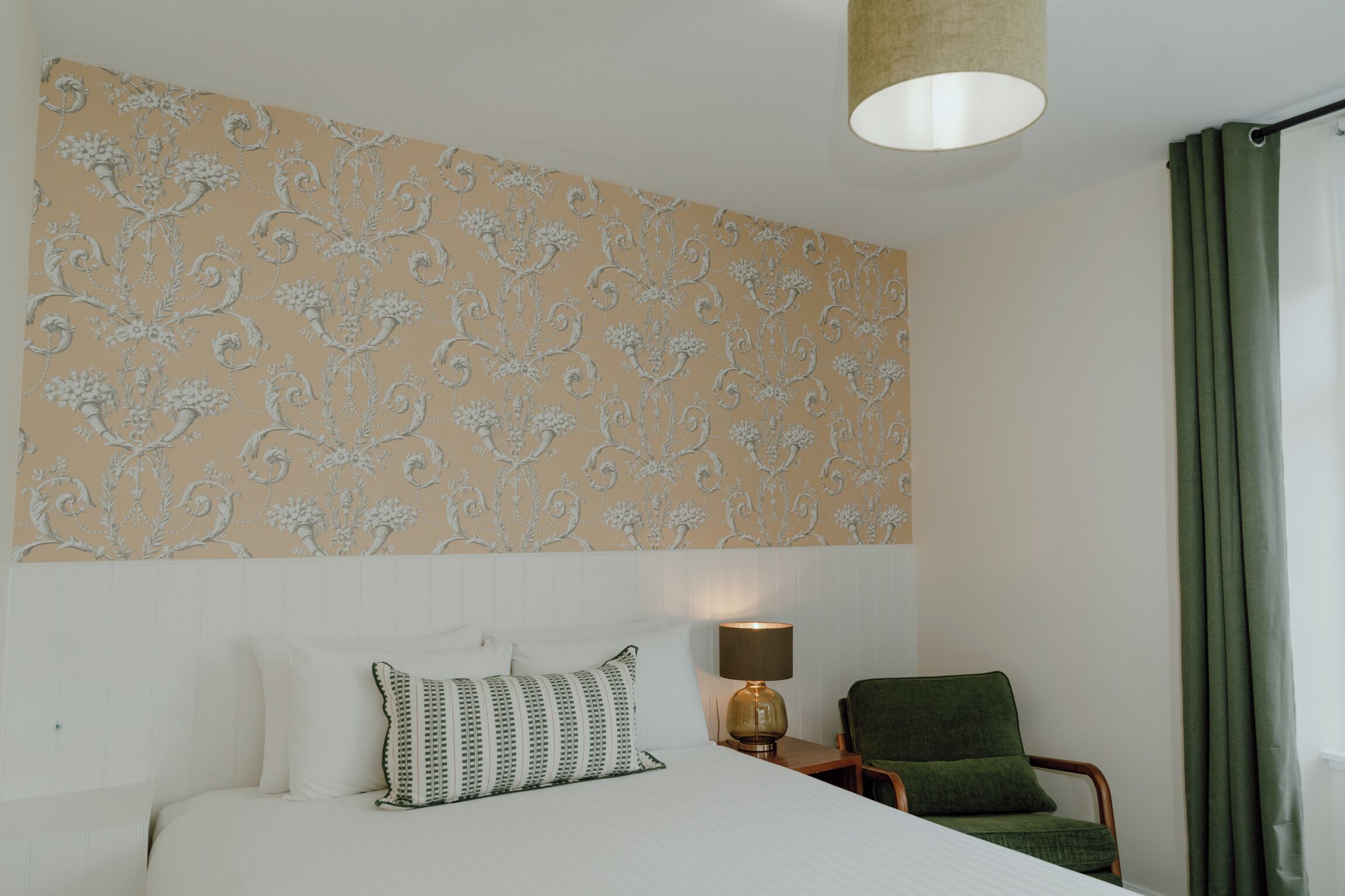 A hotel room with a bed, a chair, a lamp, and a patterned wall.