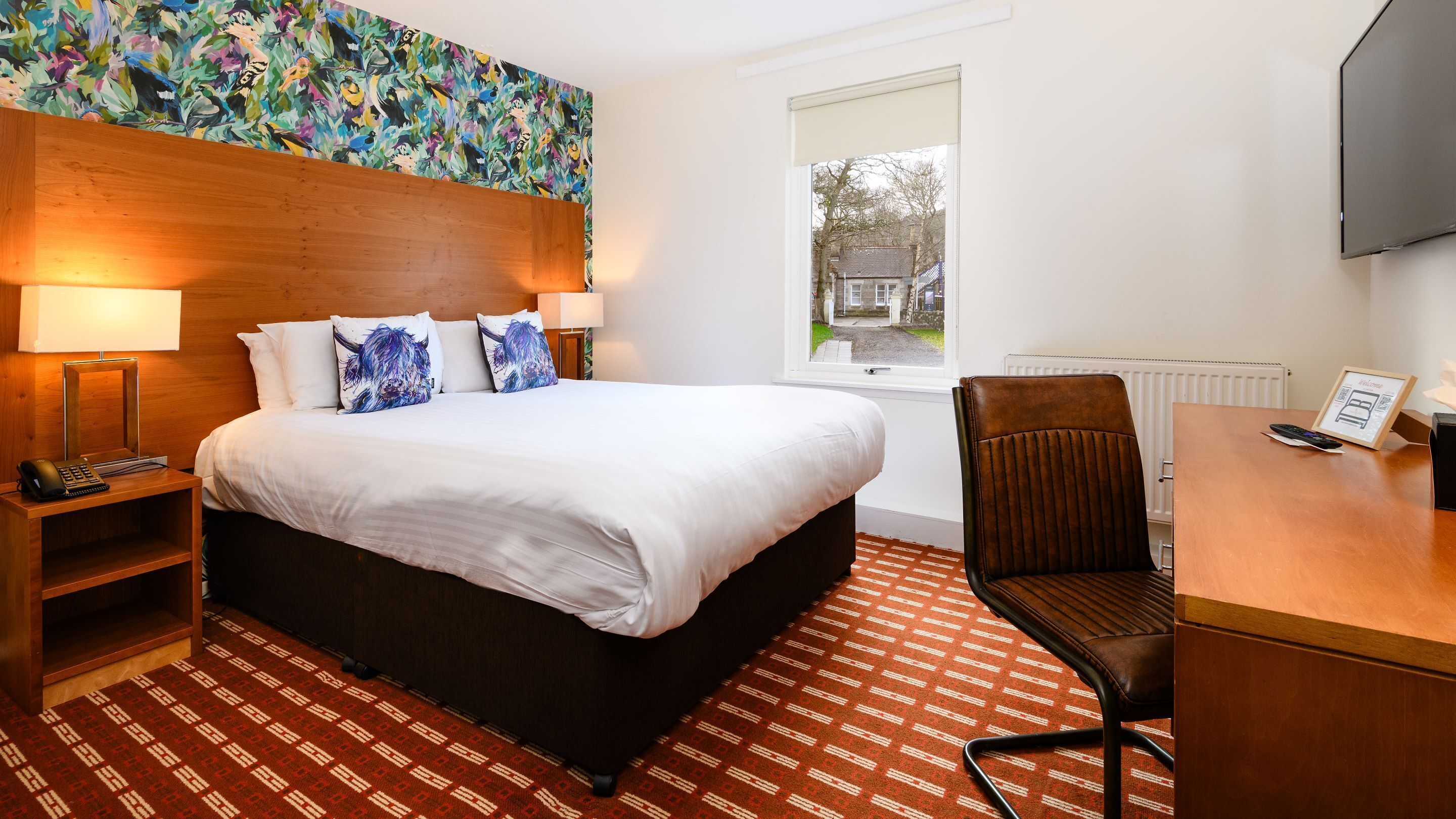 A hotel room with a large bed, two bedside lamps, a desk with a chair, a television, and a window with a view of the outside.