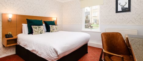 A hotel room with a large bed, a wooden headboard, a bedside table with a lamp, a red patterned carpet, and a window with a view of buildings outside.