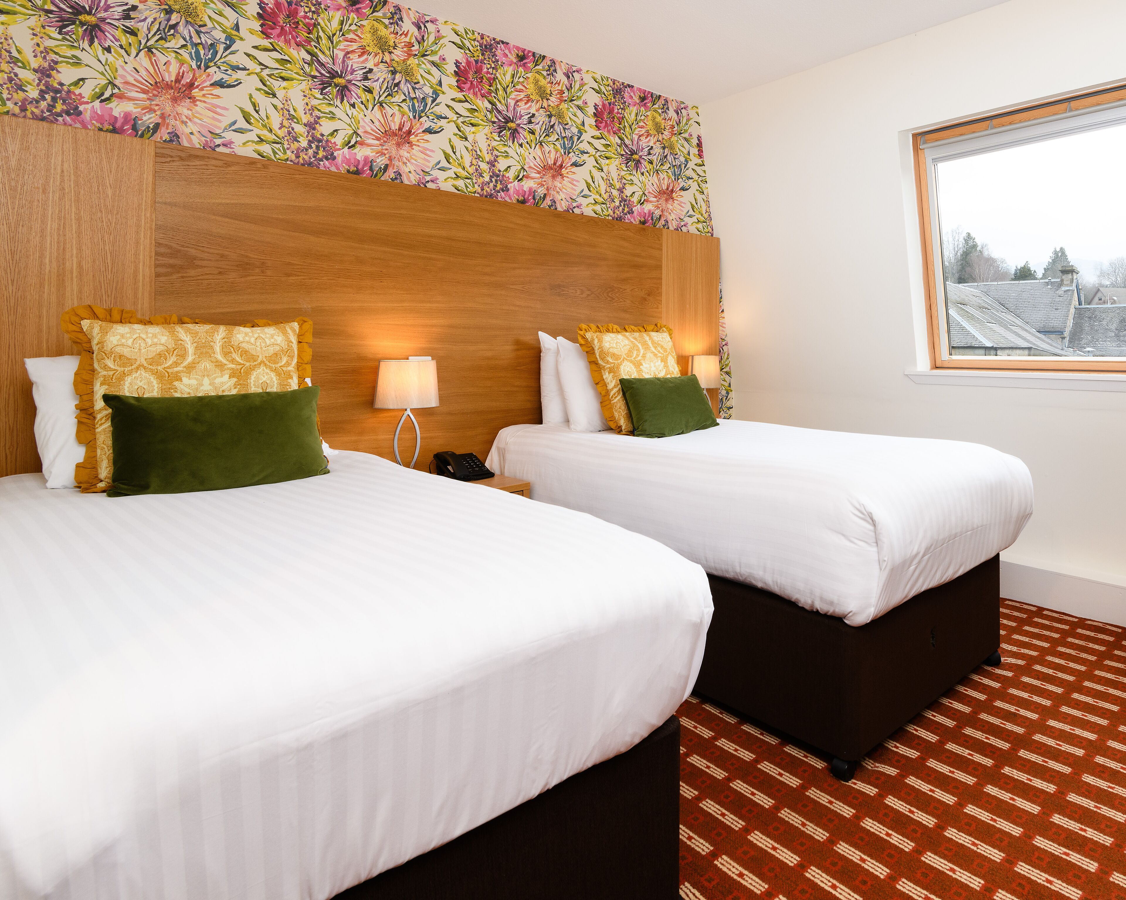 A hotel room with two beds, a wooden headboard, a floral wallpaper, a window with a view, and a patterned carpet.