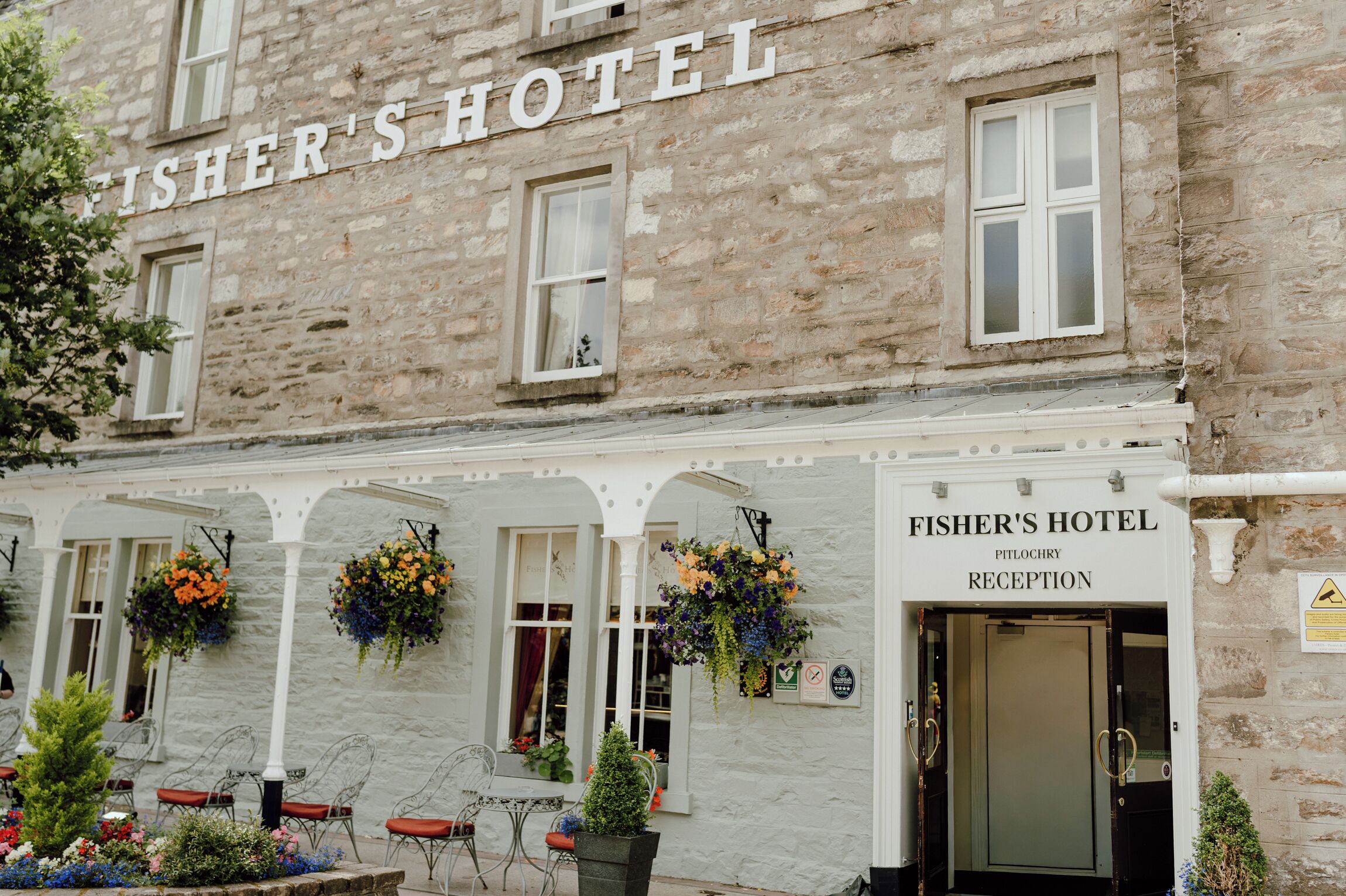 Hotel Fisher, con fachada de piedra, una zona de recepción y mesas y sillas en una terraza al aire libre.