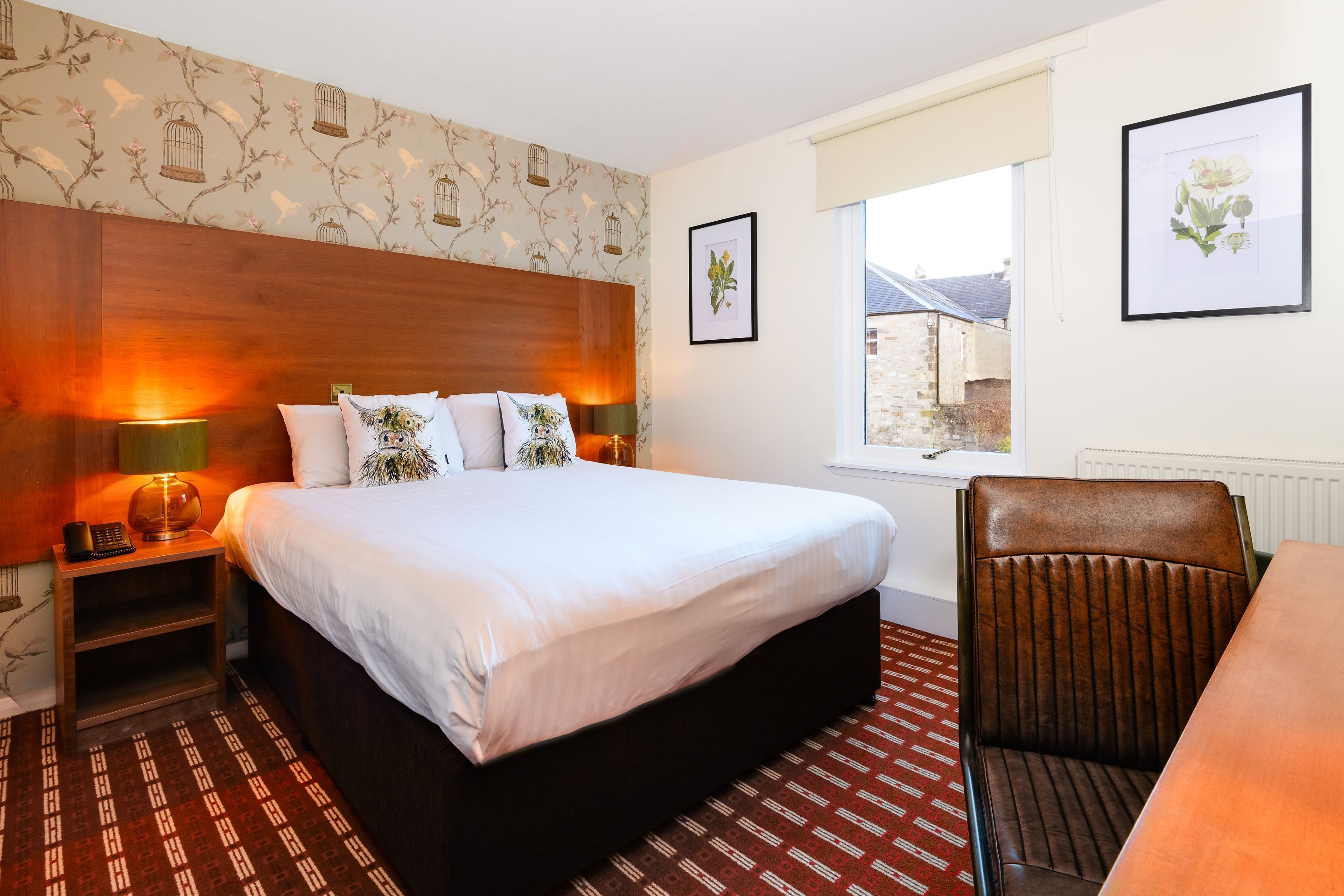 A hotel room with a large bed, a wooden headboard, a desk with a chair, and two framed botanical prints on the wall.