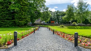 A gravel pathway flanked by flower beds and trees, leading to a building with a stone wall and a signboard. - Fisher's Hotel (Pitlochry)