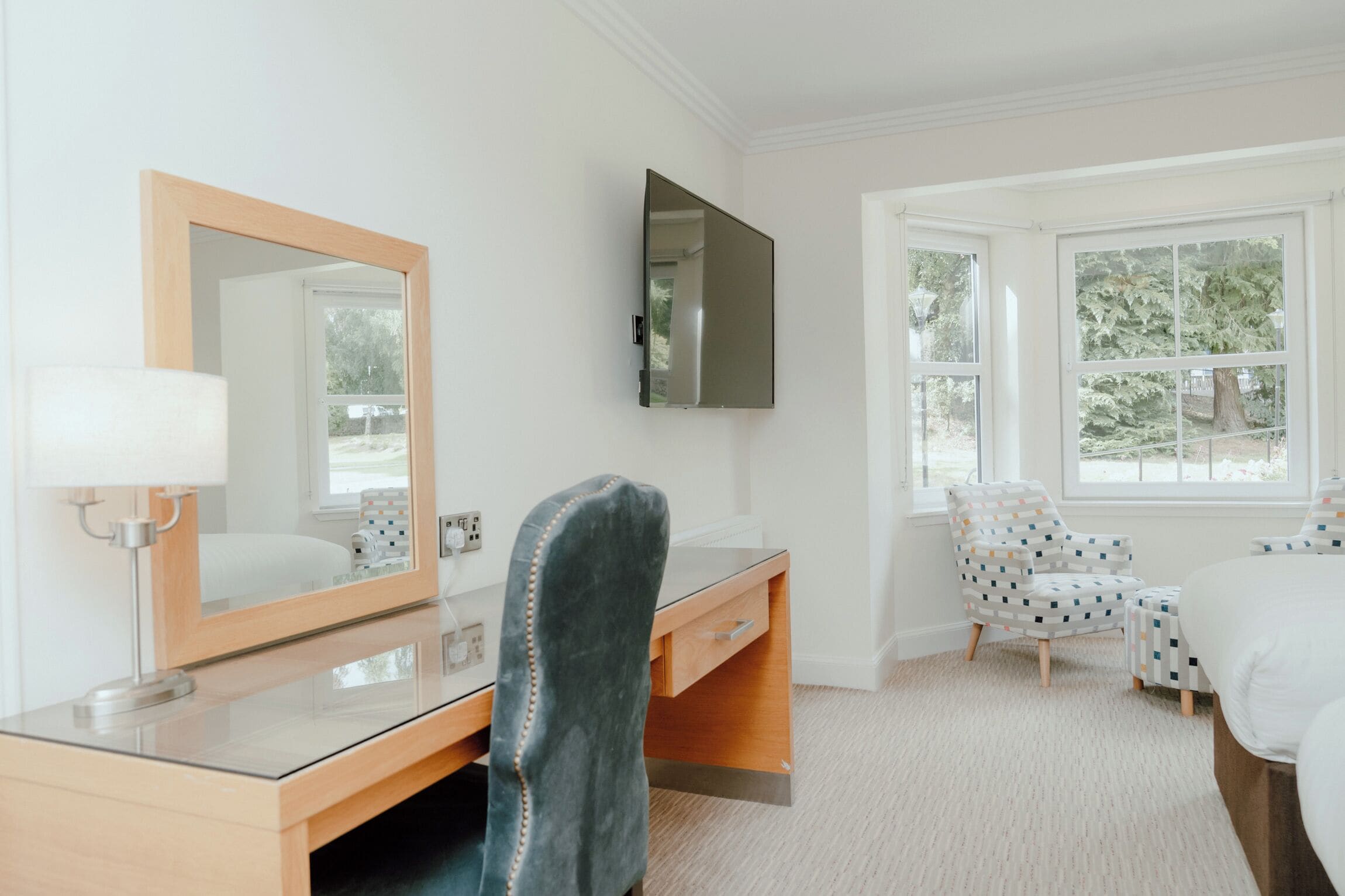 A hotel room with a wooden desk, a chair, a bed, a large mirror, and a TV mounted on the wall.