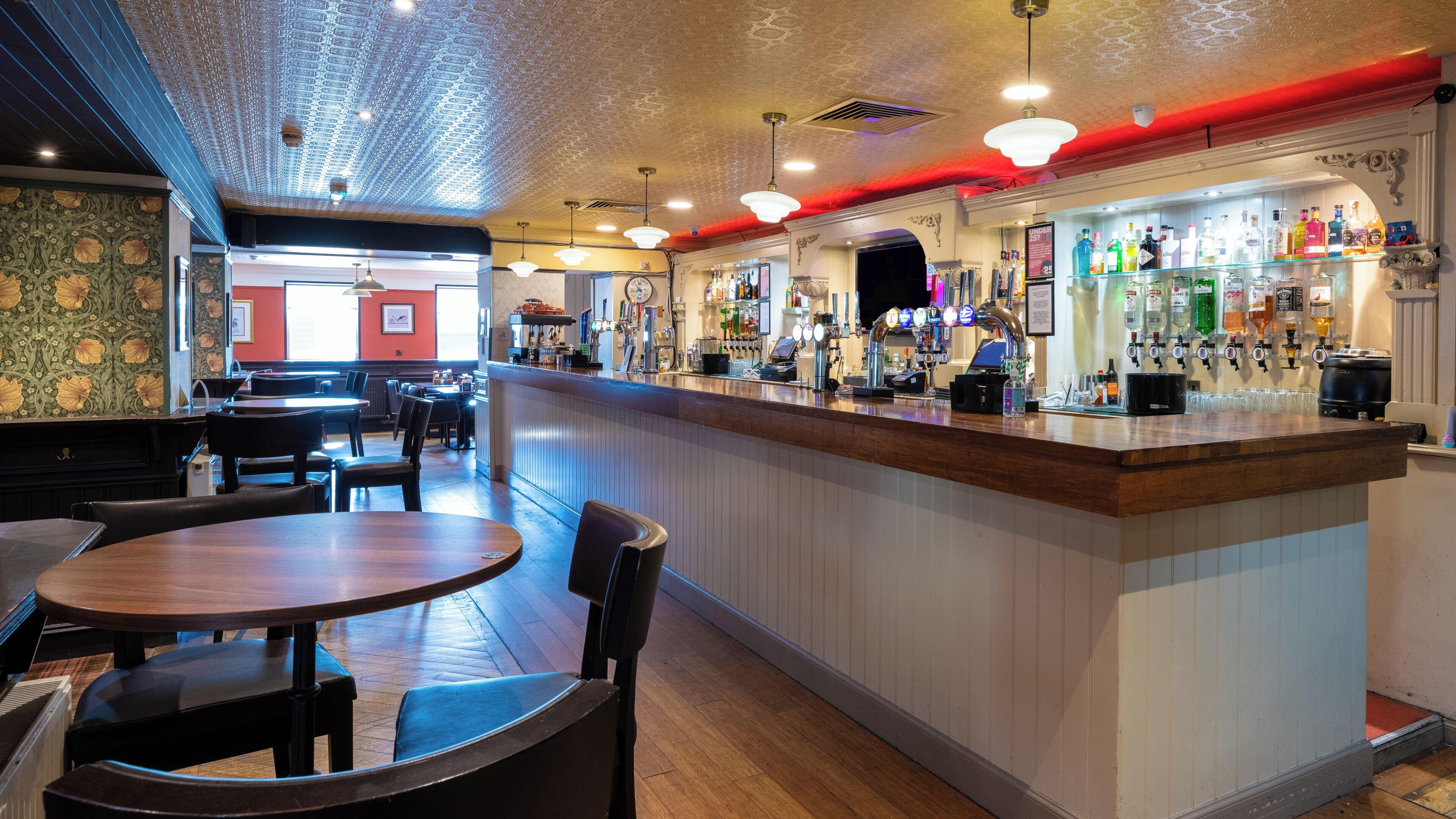 A bar area with a wooden counter, multiple taps, and a variety of bottles. There are tables and chairs for seating.