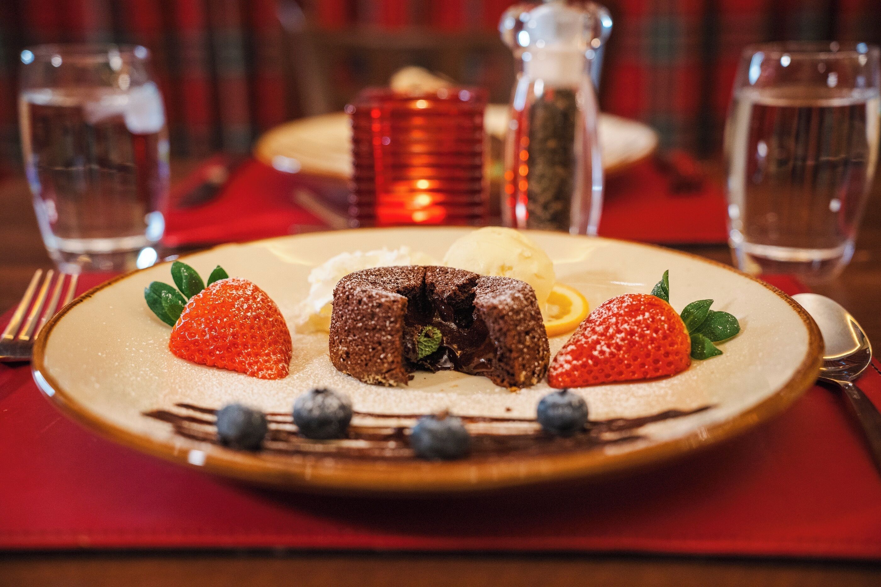 Une assiette de dessert avec des fraises, des myrtilles et un dessert au chocolat, posée sur une table avec des verres d’eau et une serviette rouge.