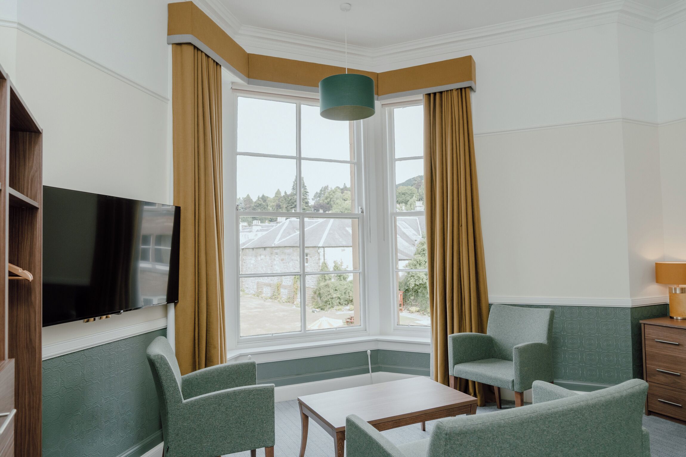 Habitación de hotel con un ventanal grande, un televisor de pantalla plana, dos sillones verdes, una mesita pequeña y un aparador de madera.