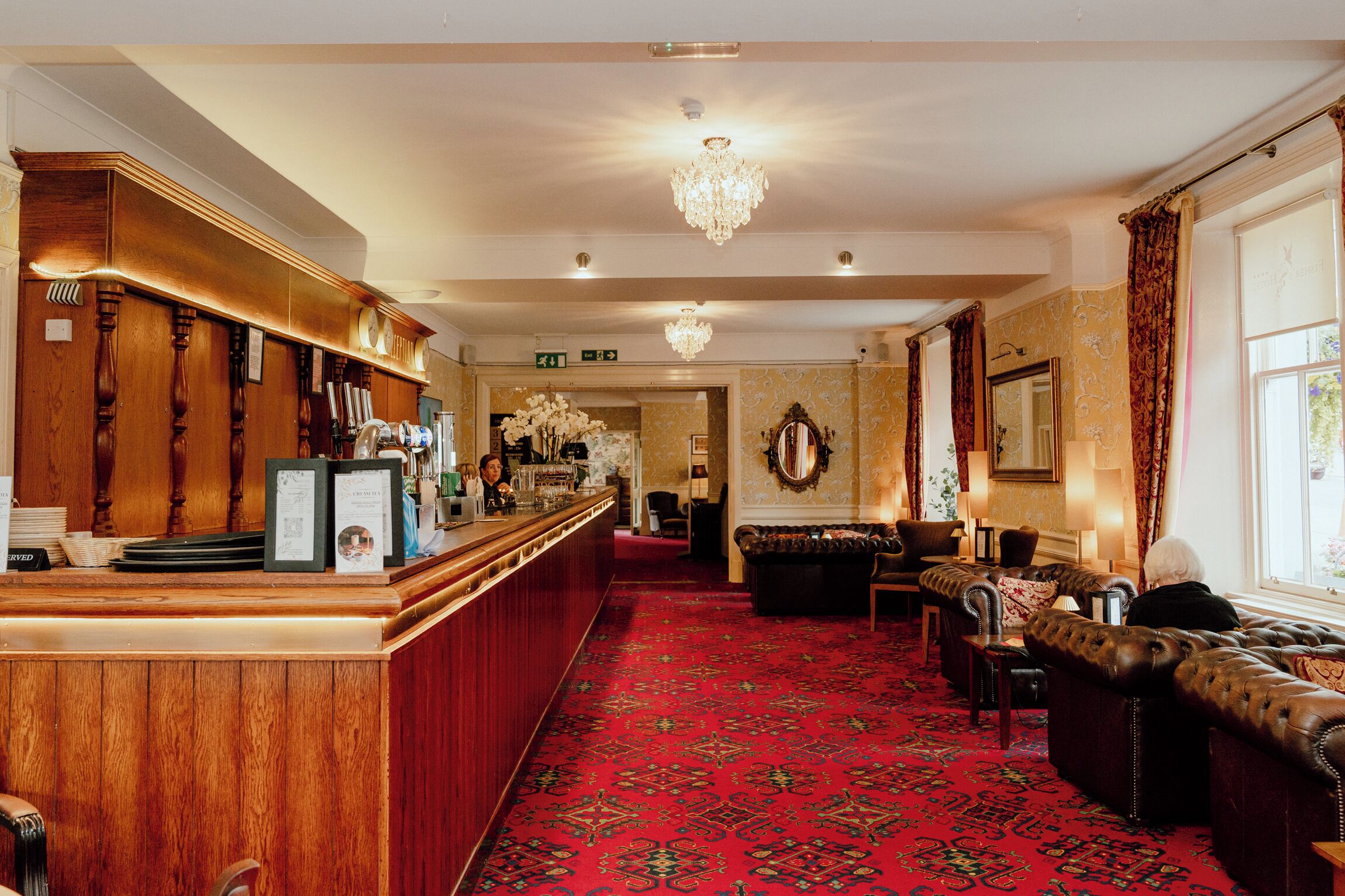 A hotel lobby with a reception desk, a chandelier, a red carpeted floor, and a seating area with leather sofas.