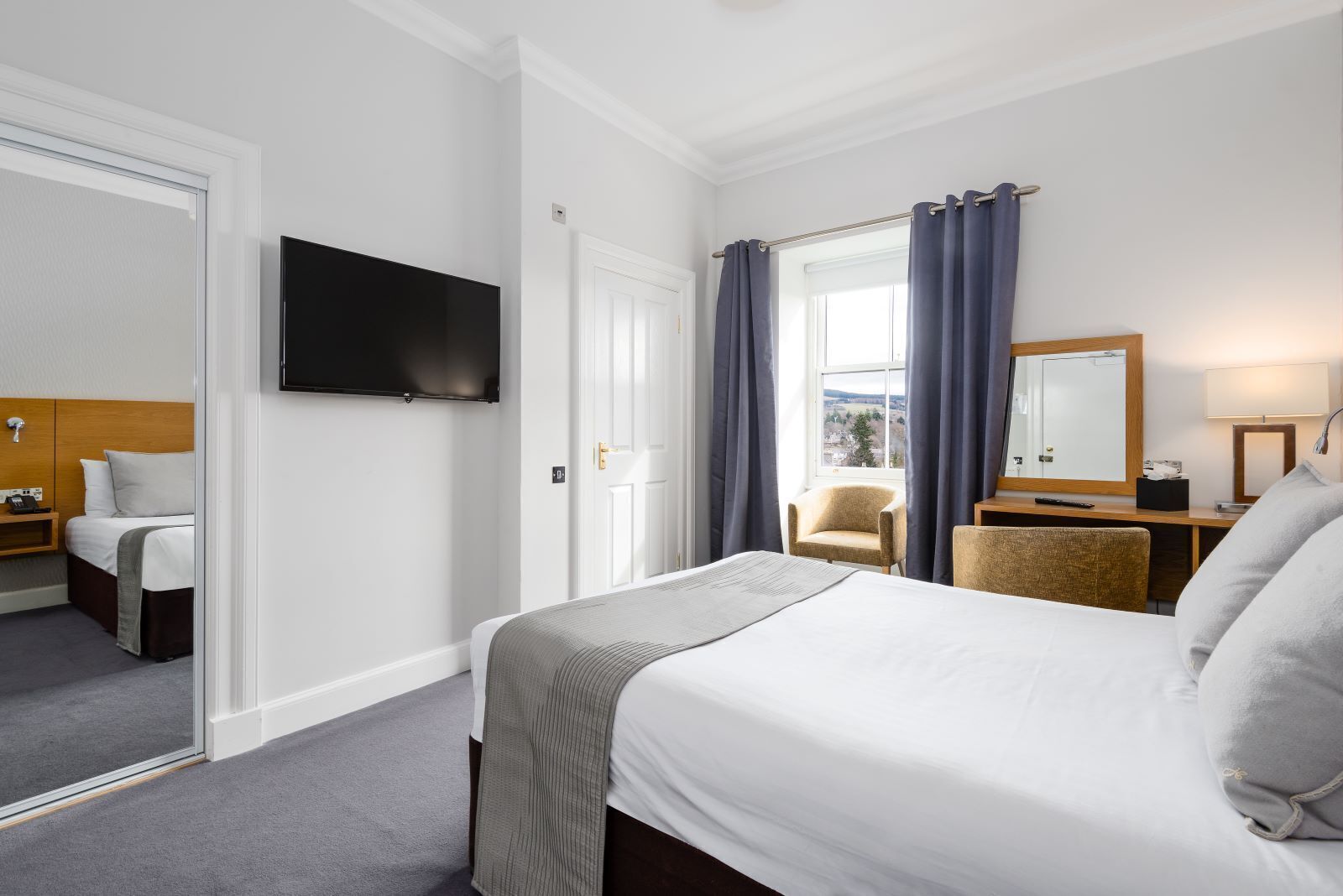 Habitación de hotel con cama, televisor, escritorio con lámpara, silla y ventana con cortinas.
