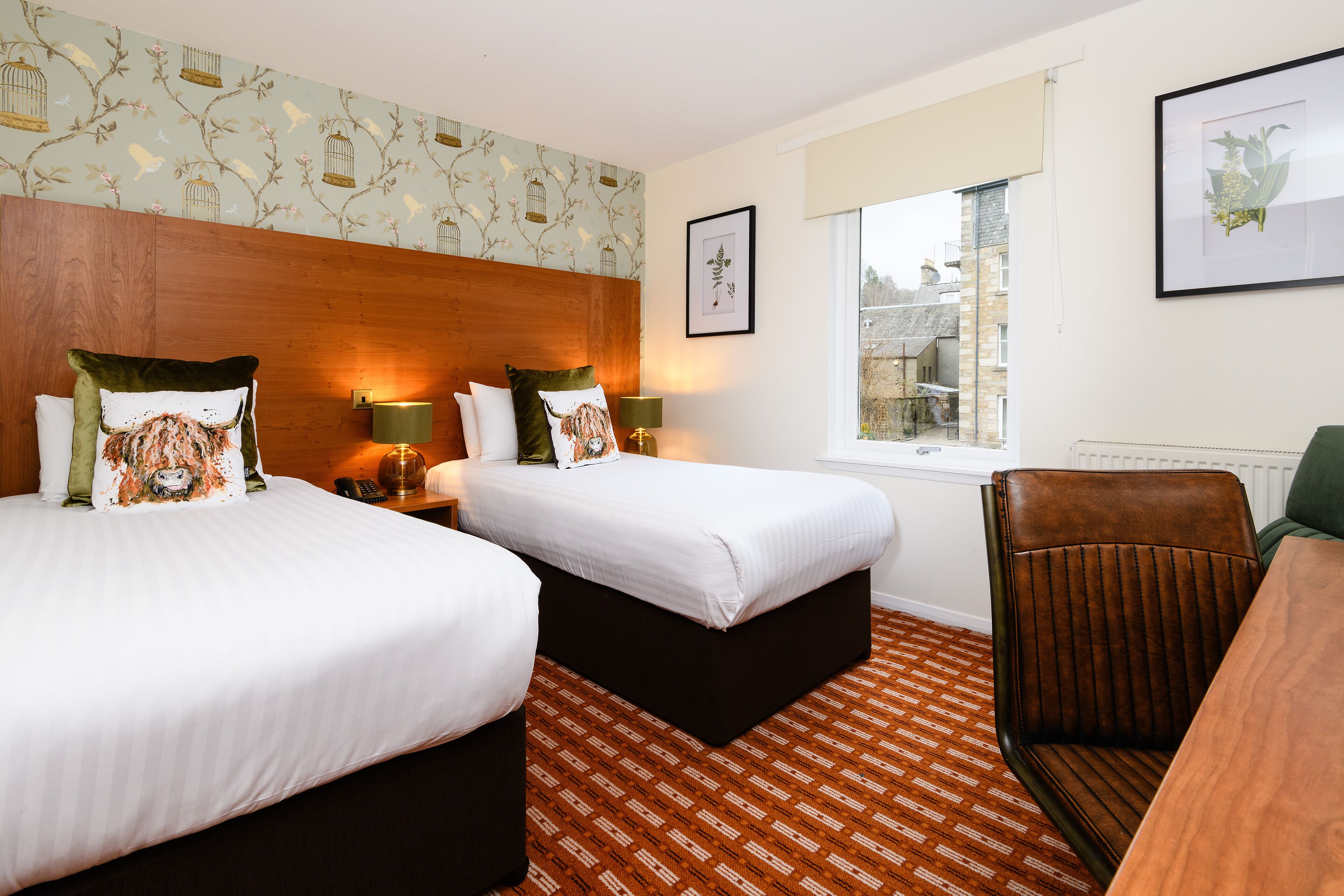 A hotel room with two beds, a wooden headboard, a window with a view, and a framed plant on the wall.