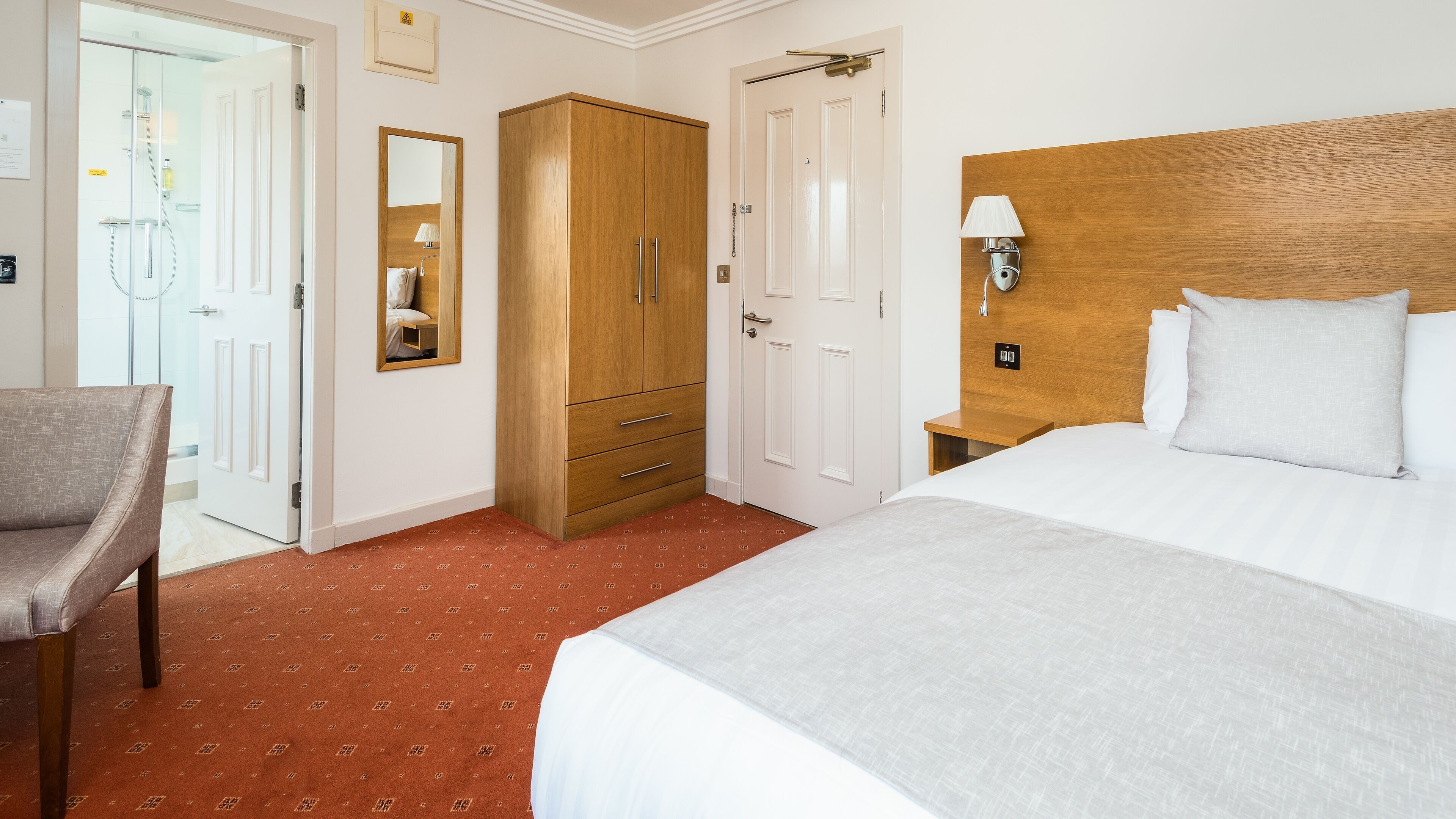 A hotel room with a bed, a wooden wardrobe, a mirror, a nightstand with a lamp, and a shower area.