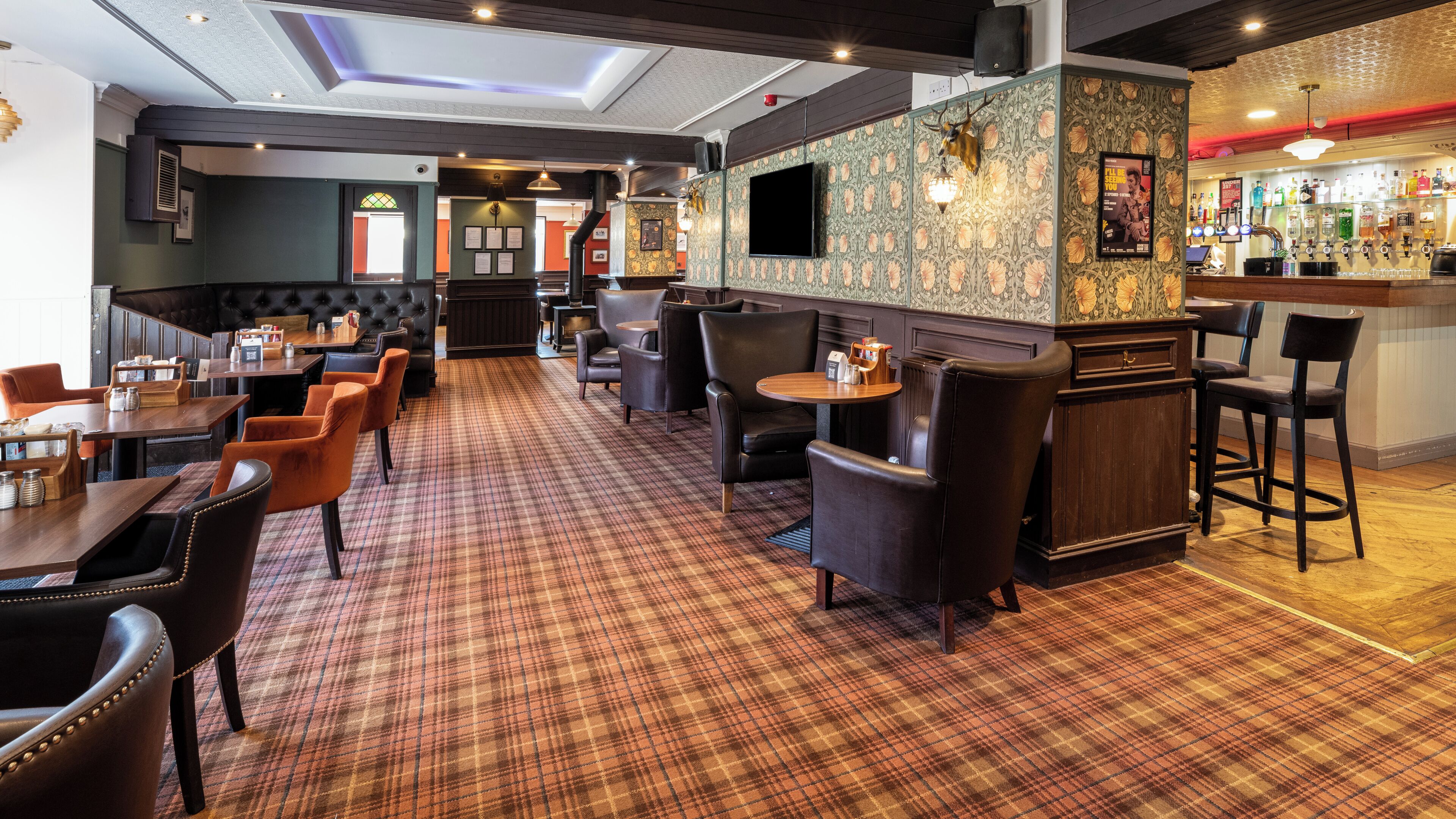A restaurant with a plaid carpet, wooden tables, leather chairs, a bar area, and a television screen.