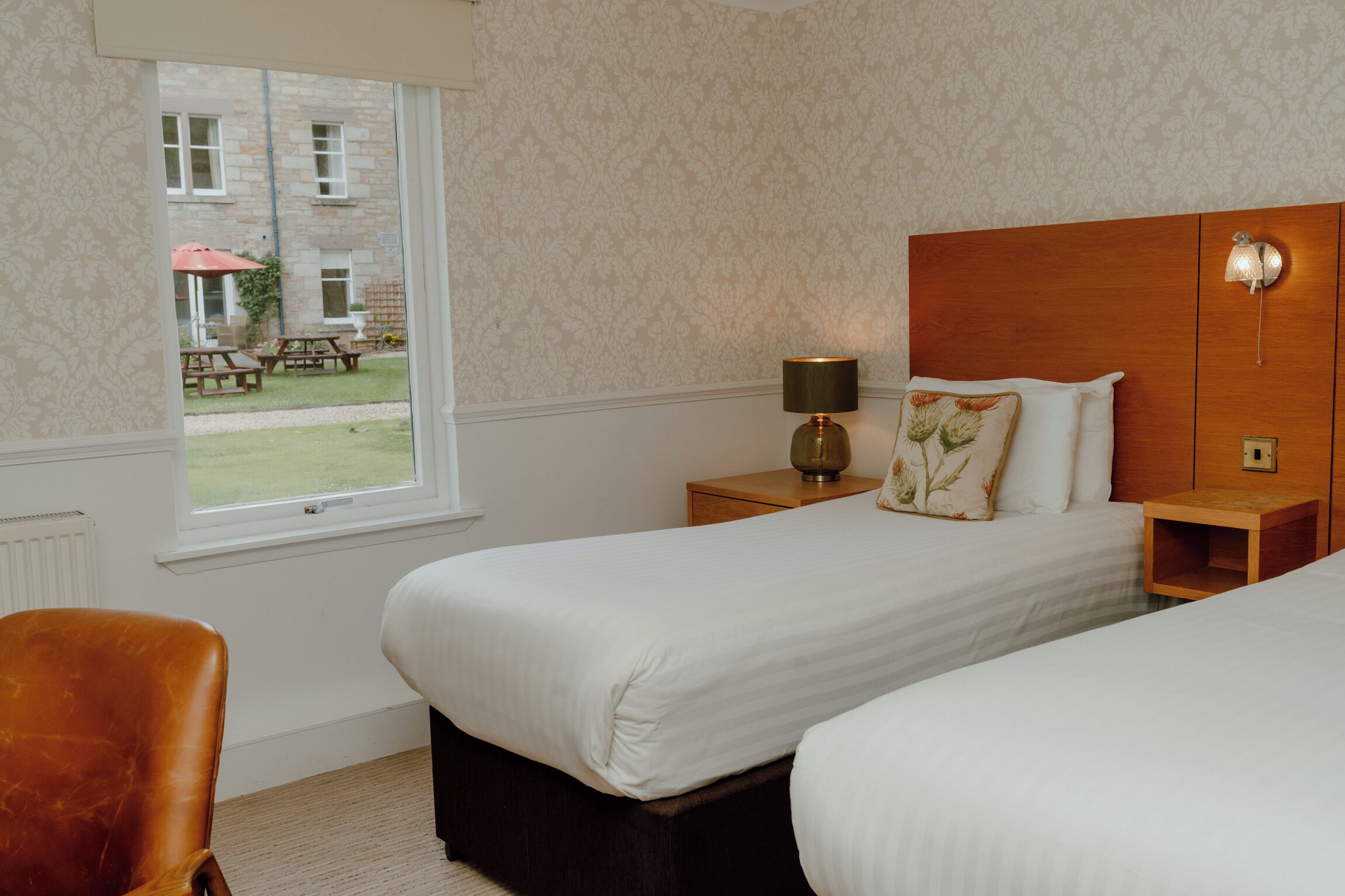 A hotel room with two beds, a wooden headboard, a nightstand with a lamp, a window with a view of the outside, and a chair.