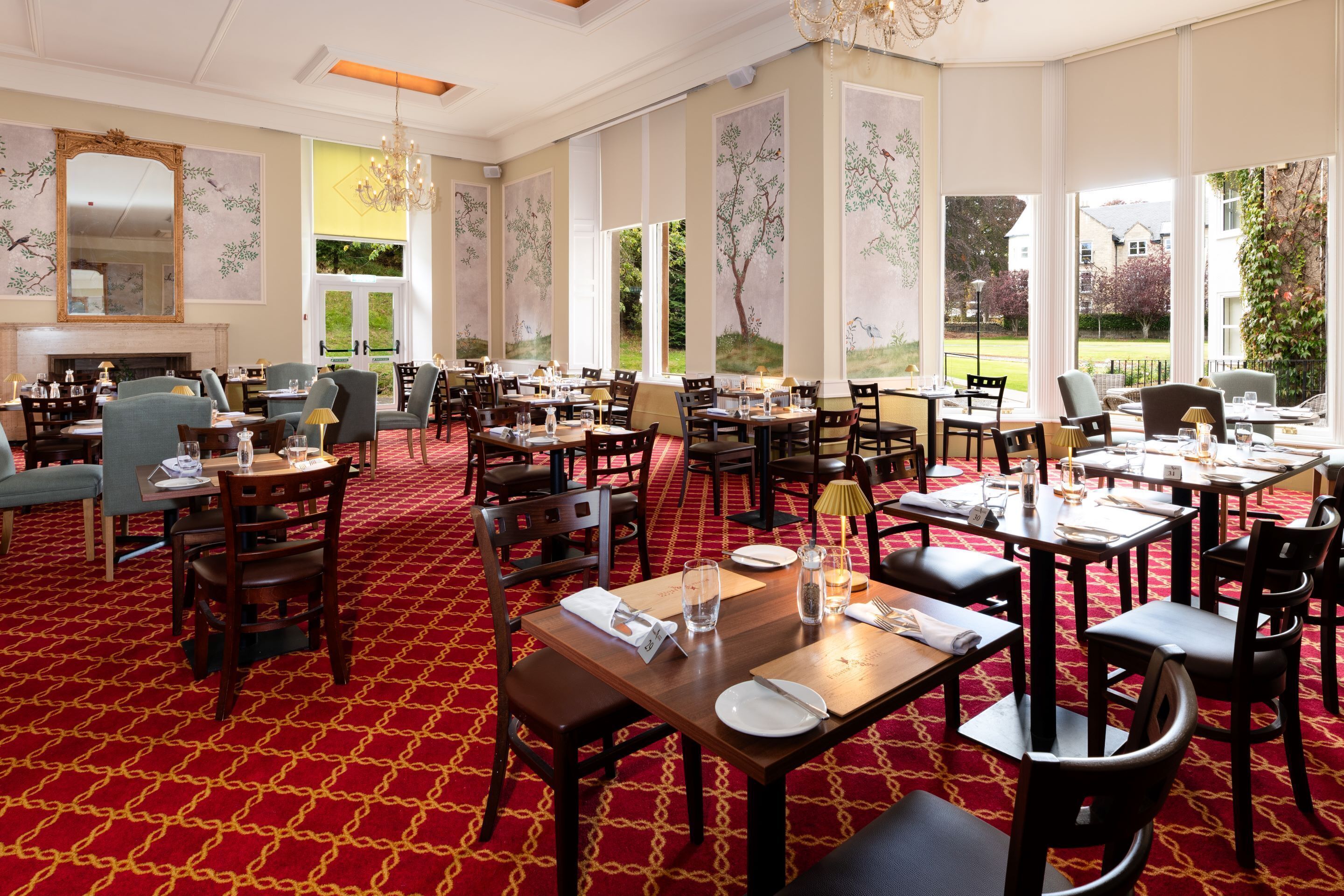 A spacious dining area with multiple tables and chairs, large windows offering views of the outdoors, and a red patterned carpet.