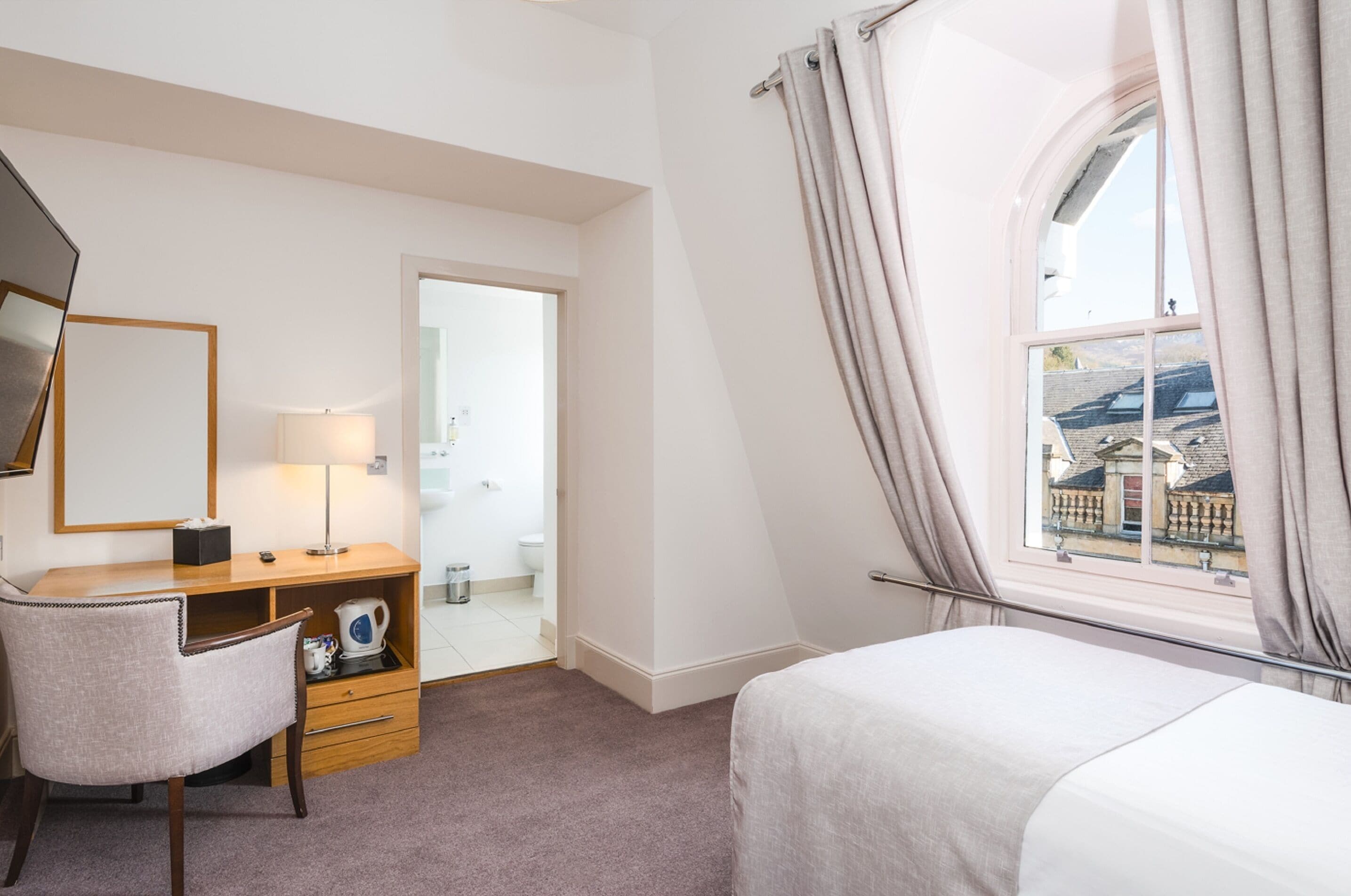 Habitación de hotel con cama, escritorio con lámpara, silla, mesita con tetera y ventana con cortinas.