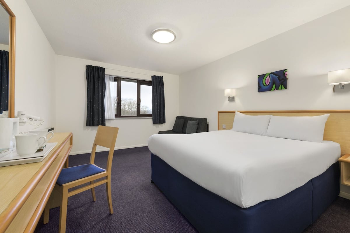 Una habitació d'hotel amb un llit gran, una taula de treball amb una cadira, una finestra amb cortines, un quadre a la paret i una tauleta de nit.